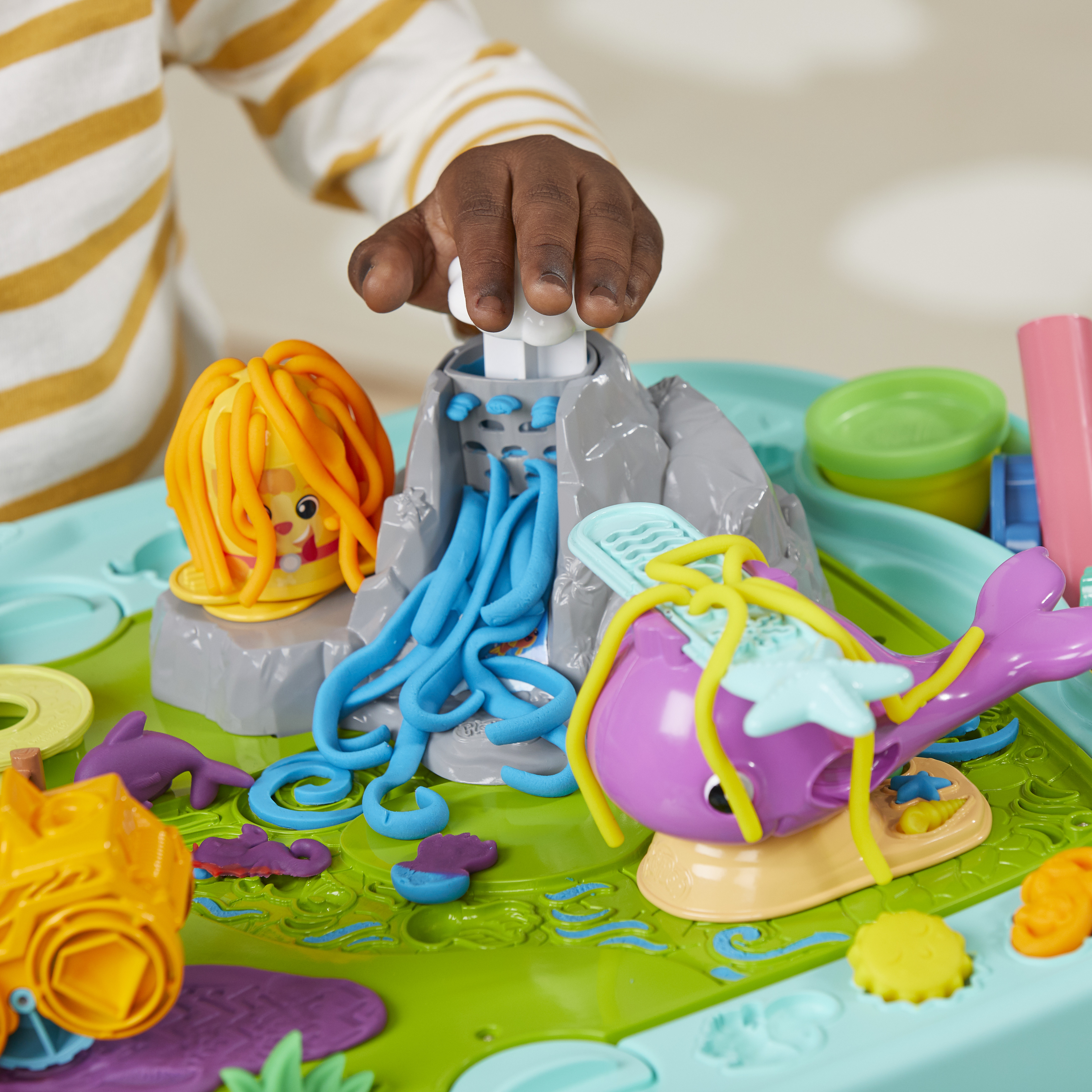 La mano de un niño interactúa con un set de Play-Doh. Presenta un volcán, una sirena, una ballena y varias formas coloridas de Play-Doh.