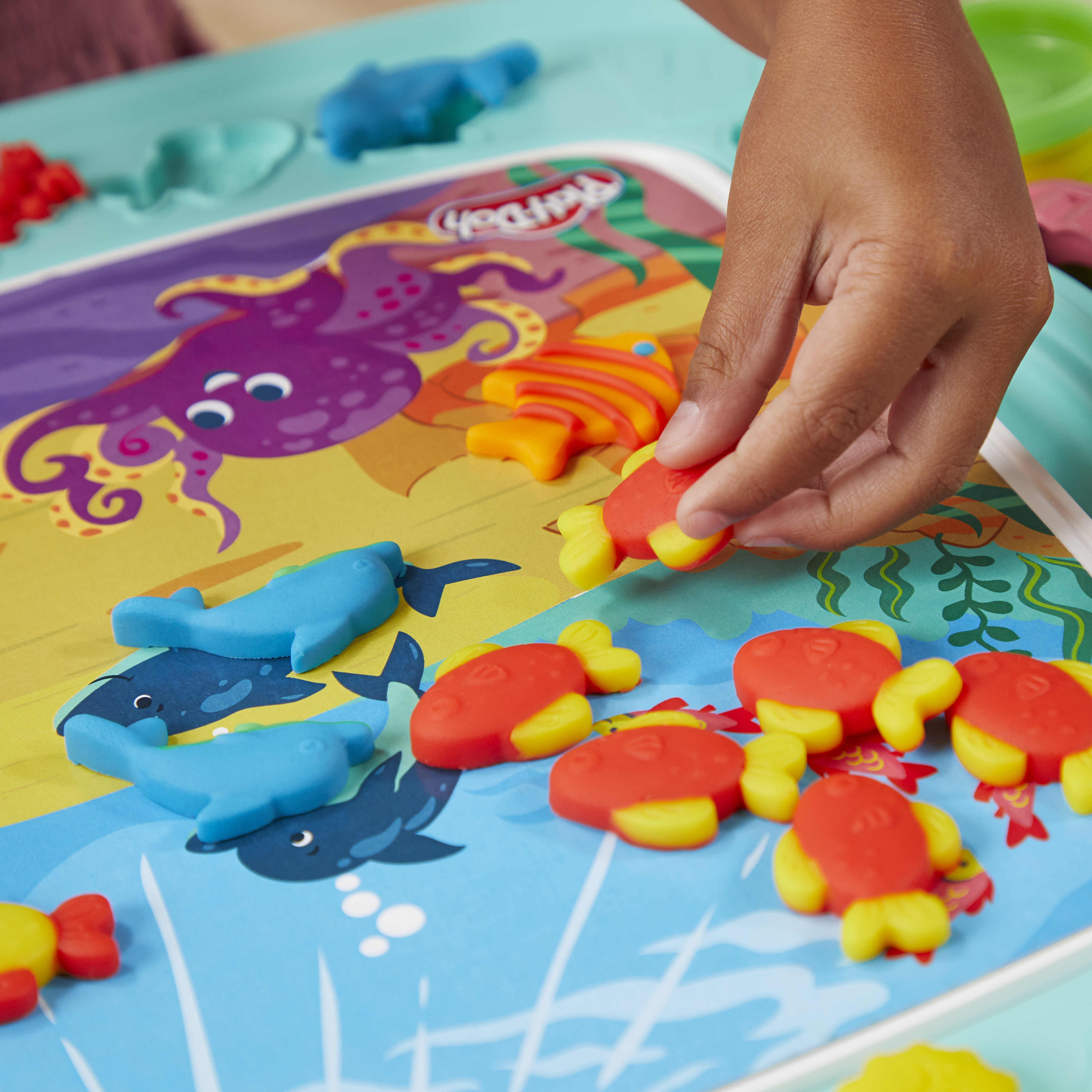 Una mano colocando un pez rojo-amarillo en un tapete de juego Play-Doh con figuras de ballenas azules.