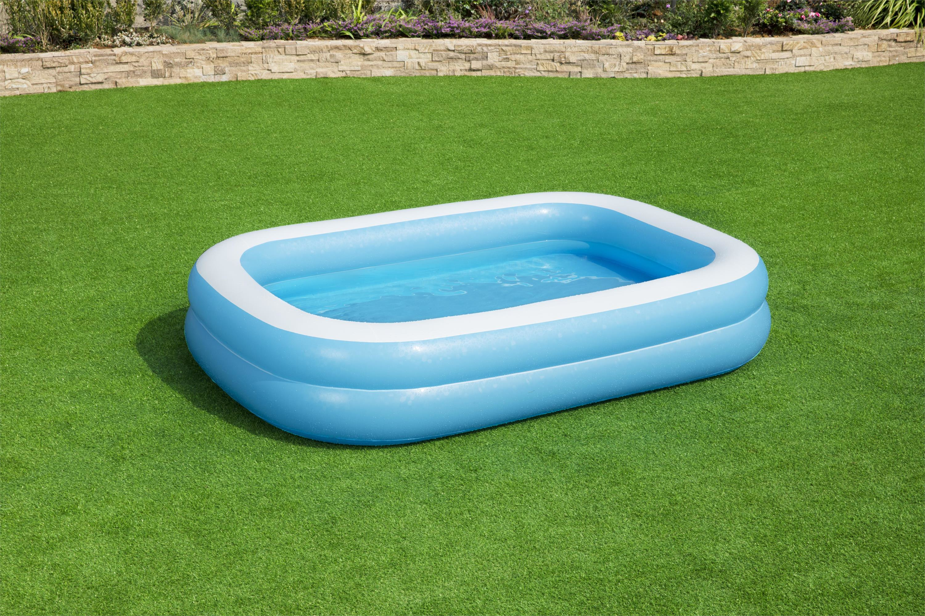 Una piscina inflable azul llena de agua se encuentra sobre césped verde. Al fondo, una pared de piedra con plantas.