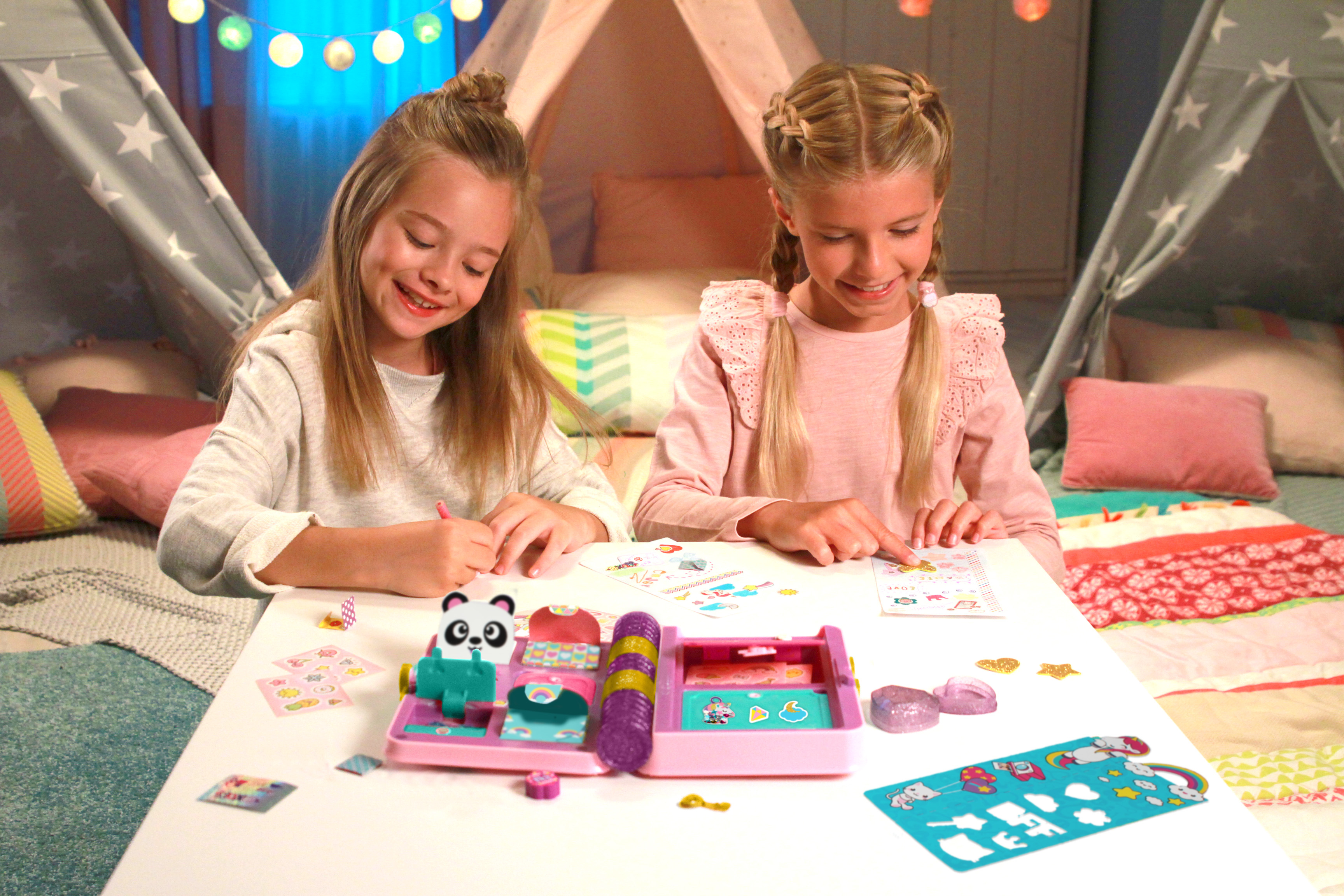 Dwie dziewczyny siedzą przy stole i tworzą. Zabawka z pandą, naklejki i namiot w tle.