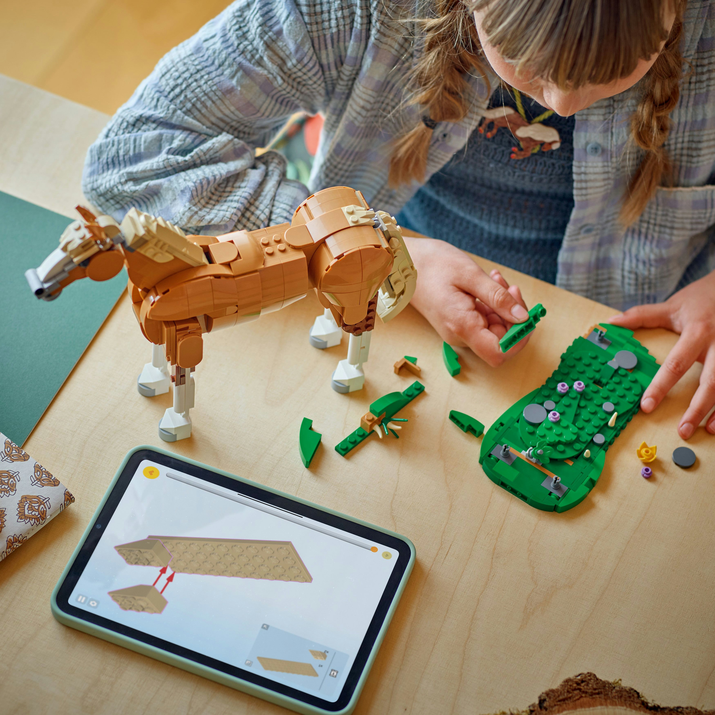 Dziewczyna buduje konia i krajobraz z klocków Lego. Tablet pokazuje instrukcje. Drewniany stół. Widoczne są brązowe, beżowe i zielone klocki Lego.