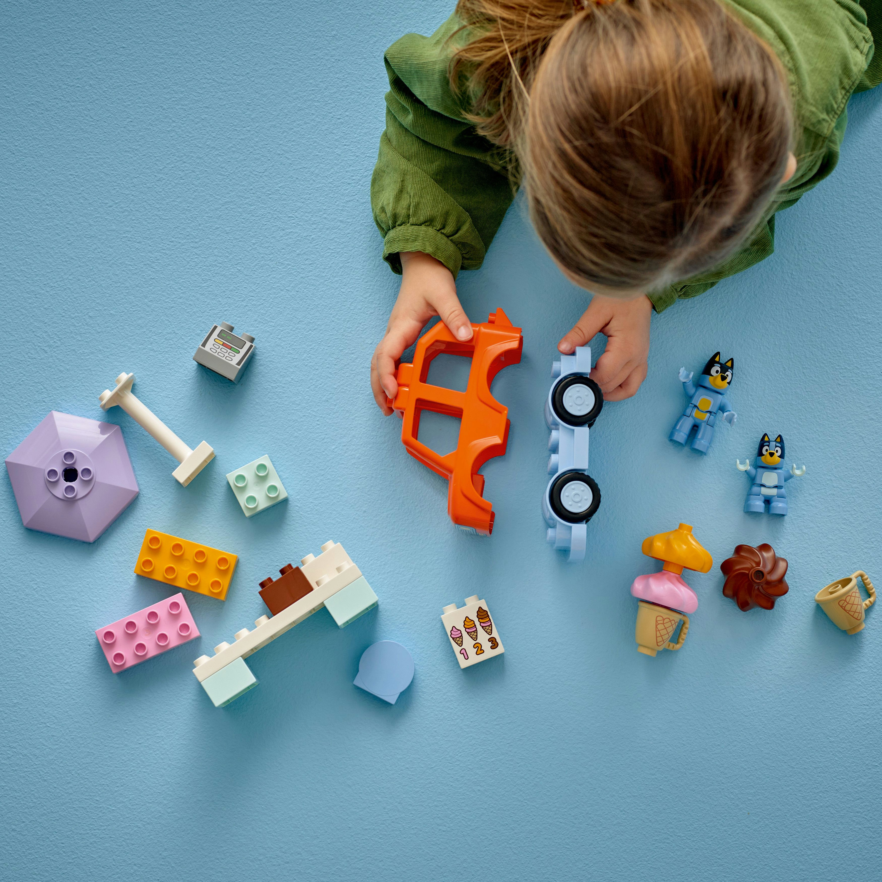 Dziecko bawi się klockami LEGO Duplo. Na niebieskiej powierzchni znajdują się części samochodów, figurki Bluey, klocki i rożki do lodów.