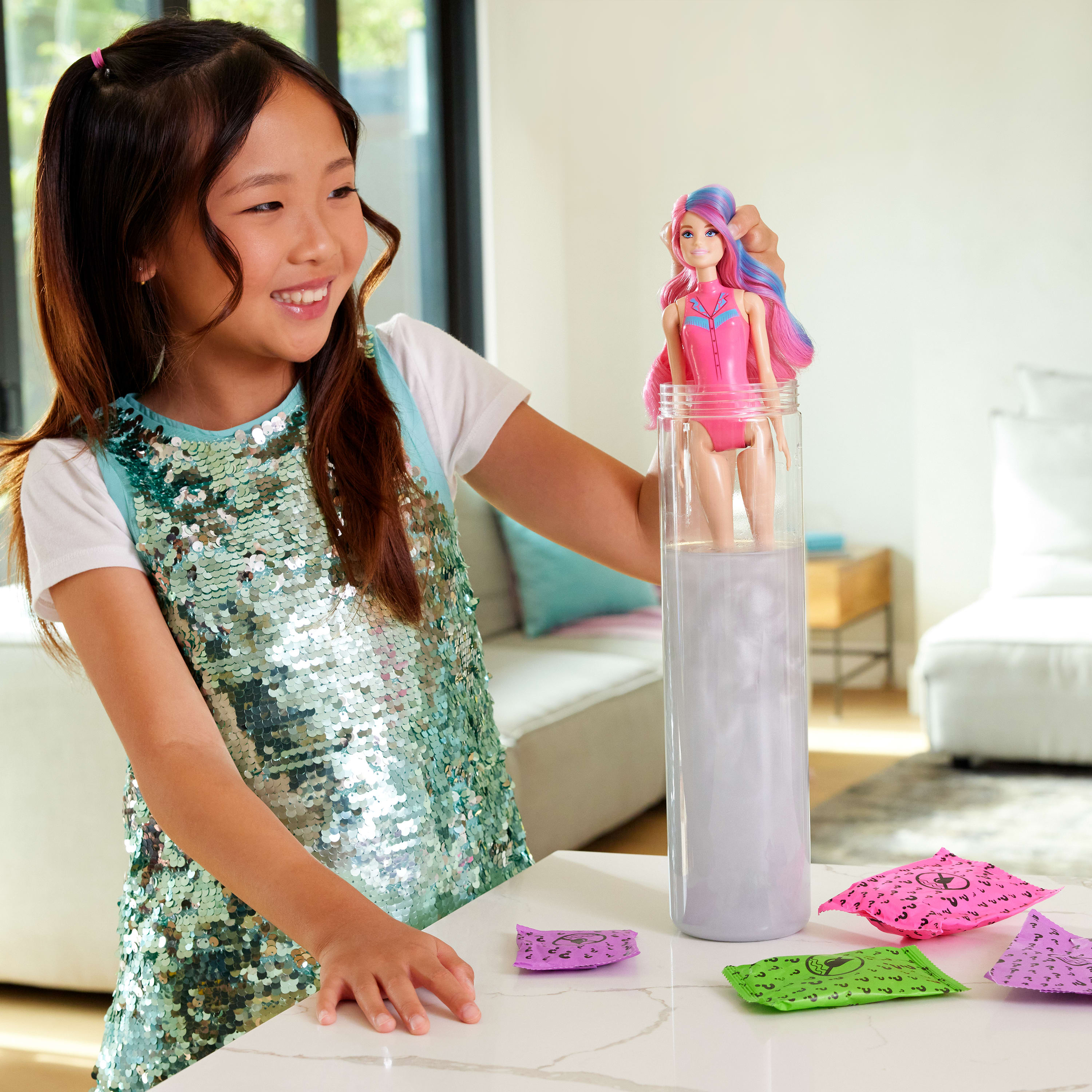 Una niña sostiene una muñeca en un recipiente. La muñeca tiene un atuendo rosa y cabello colorido. Varios paquetes de colores sobre la mesa.