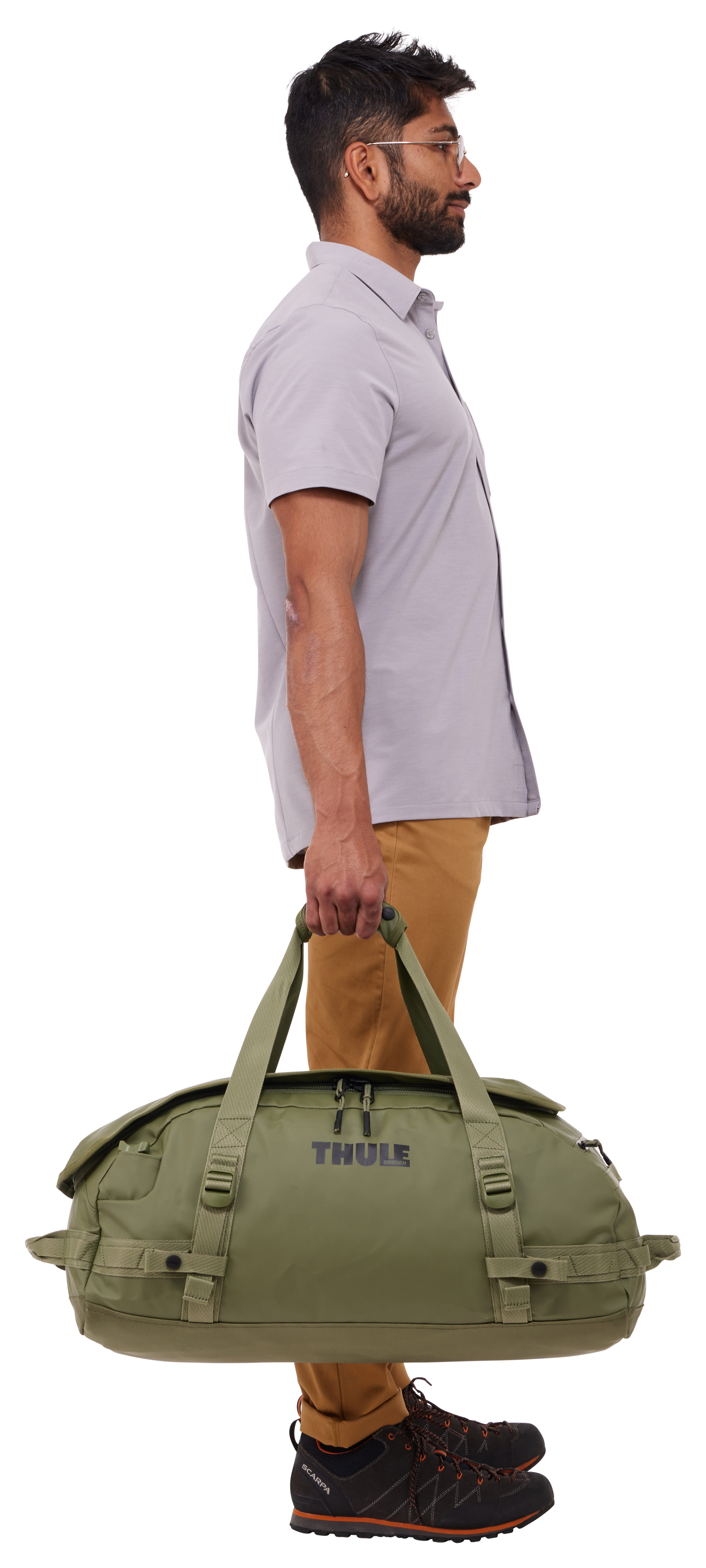 Mężczyzna w jasnej koszuli i spodniach trzyma zieloną torbę podróżną. Ma okulary i buty trekkingowe.