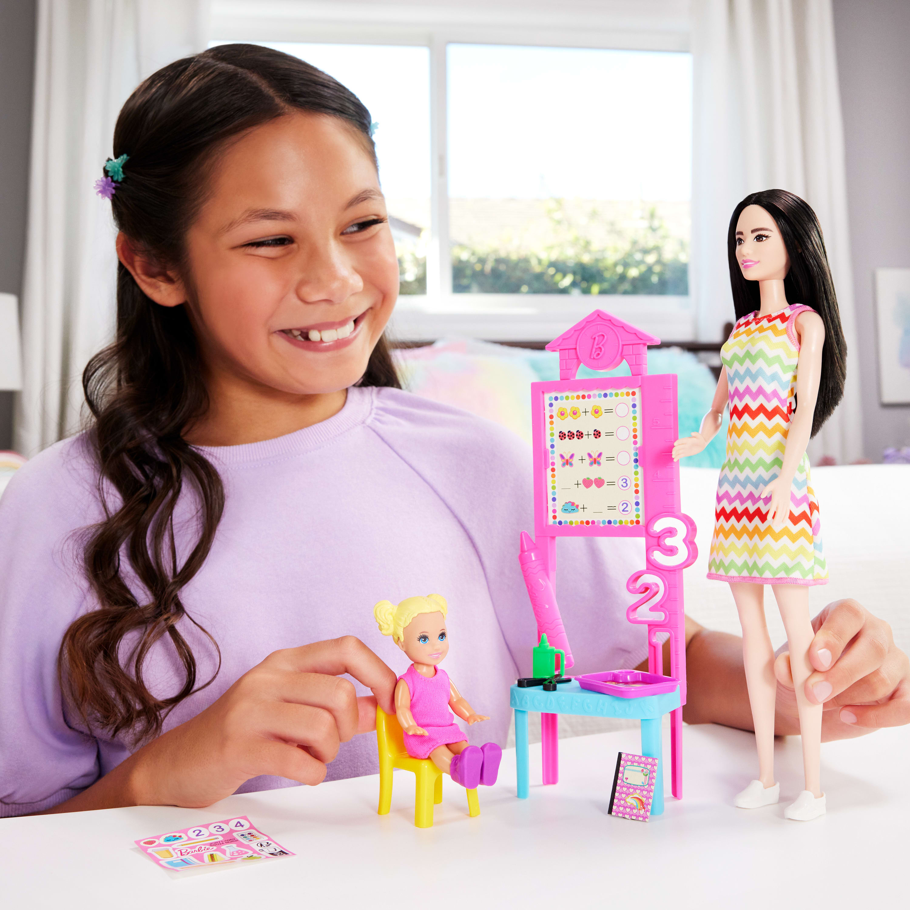 Ein lächelndes Mädchen spielt mit Barbie-Puppen und Zubehör. Eine Puppe steht, eine andere sitzt auf einem gelben Stuhl.