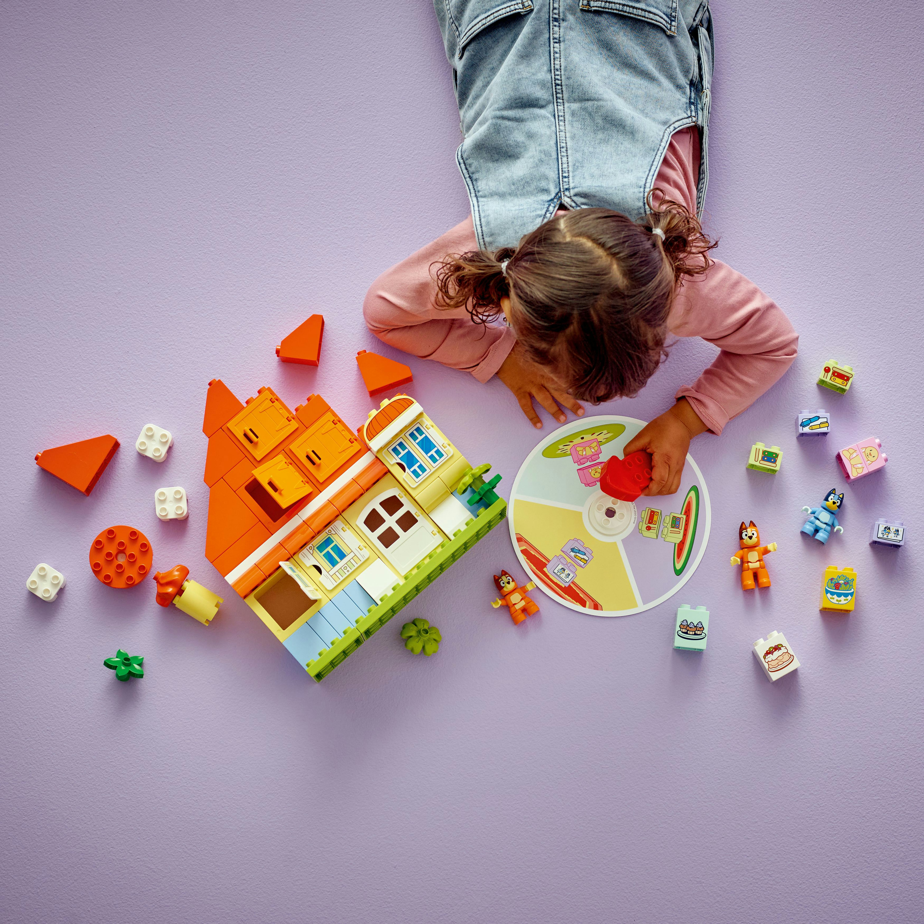 Dziecko bawi się klockami na fioletowej powierzchni. Widoczny jest dom, figurki i rozrzucone klocki.