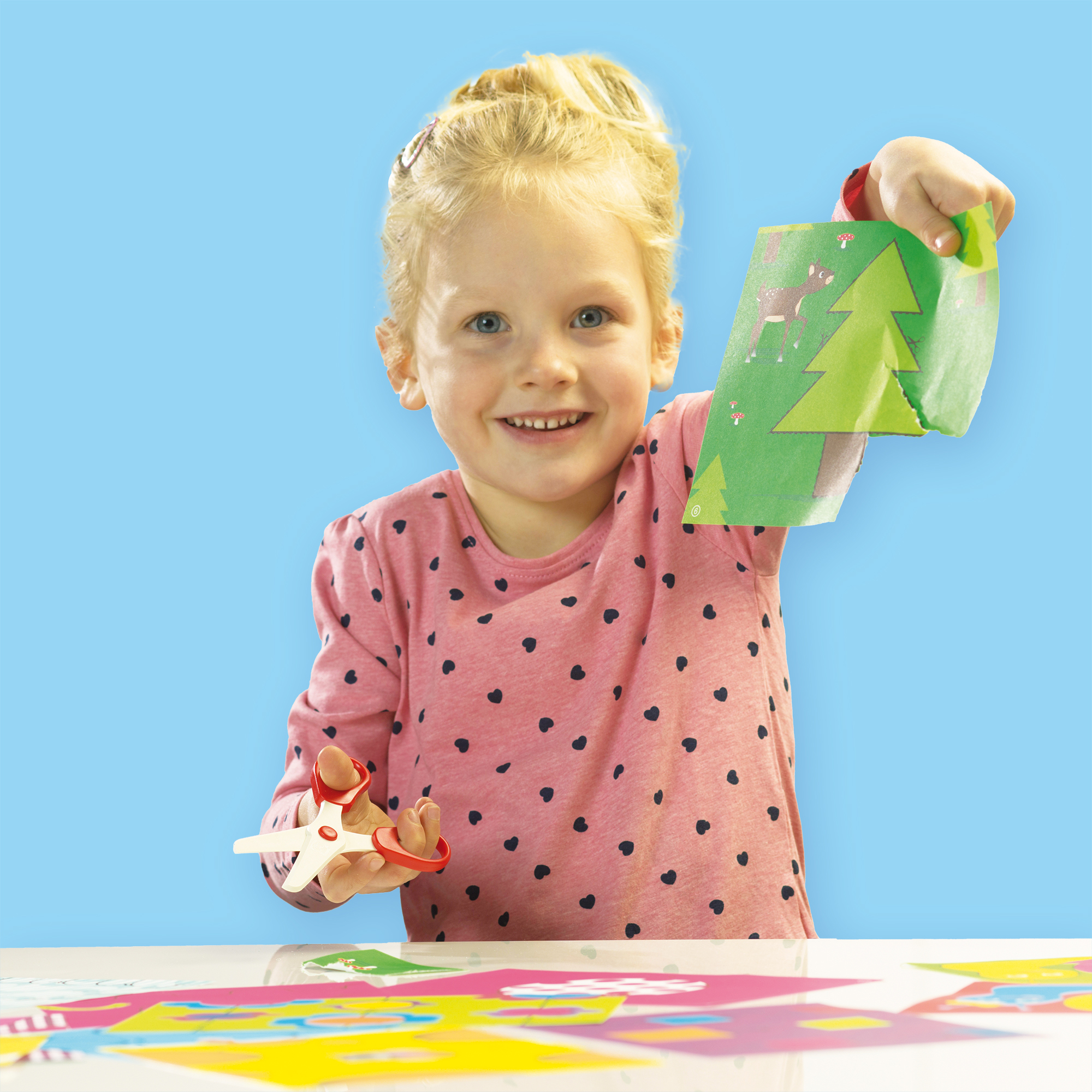 Ein lächelndes blondes Mädchen hält ein grünes Papier mit Hirsch- und Baumdesign, Schere in der Hand, auf blauem Hintergrund.