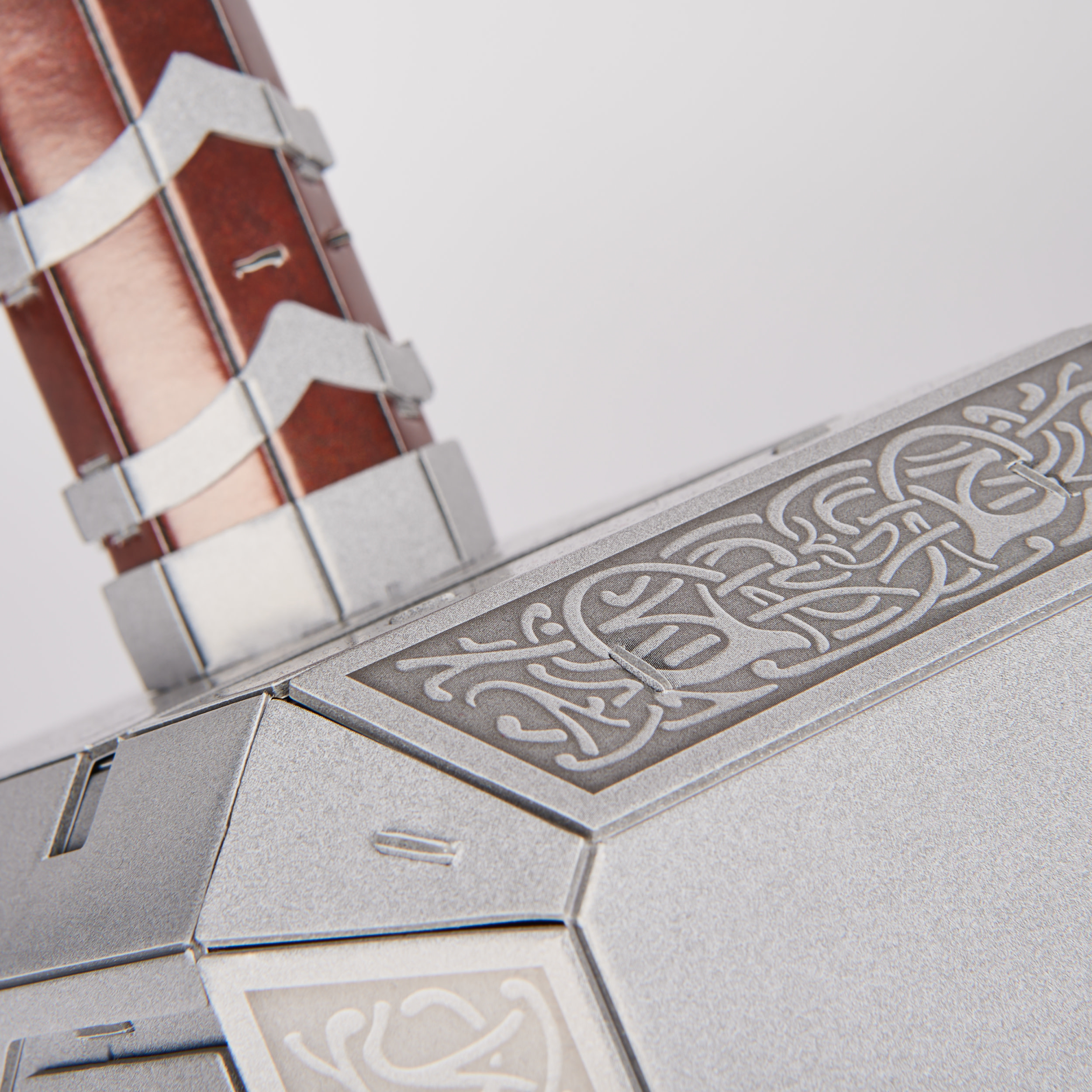 Zbliżenie młota Thora, Mjolnira. Srebrna metalowa głowica z grawerowanym wzorem, brązowy uchwyt i białe tło.