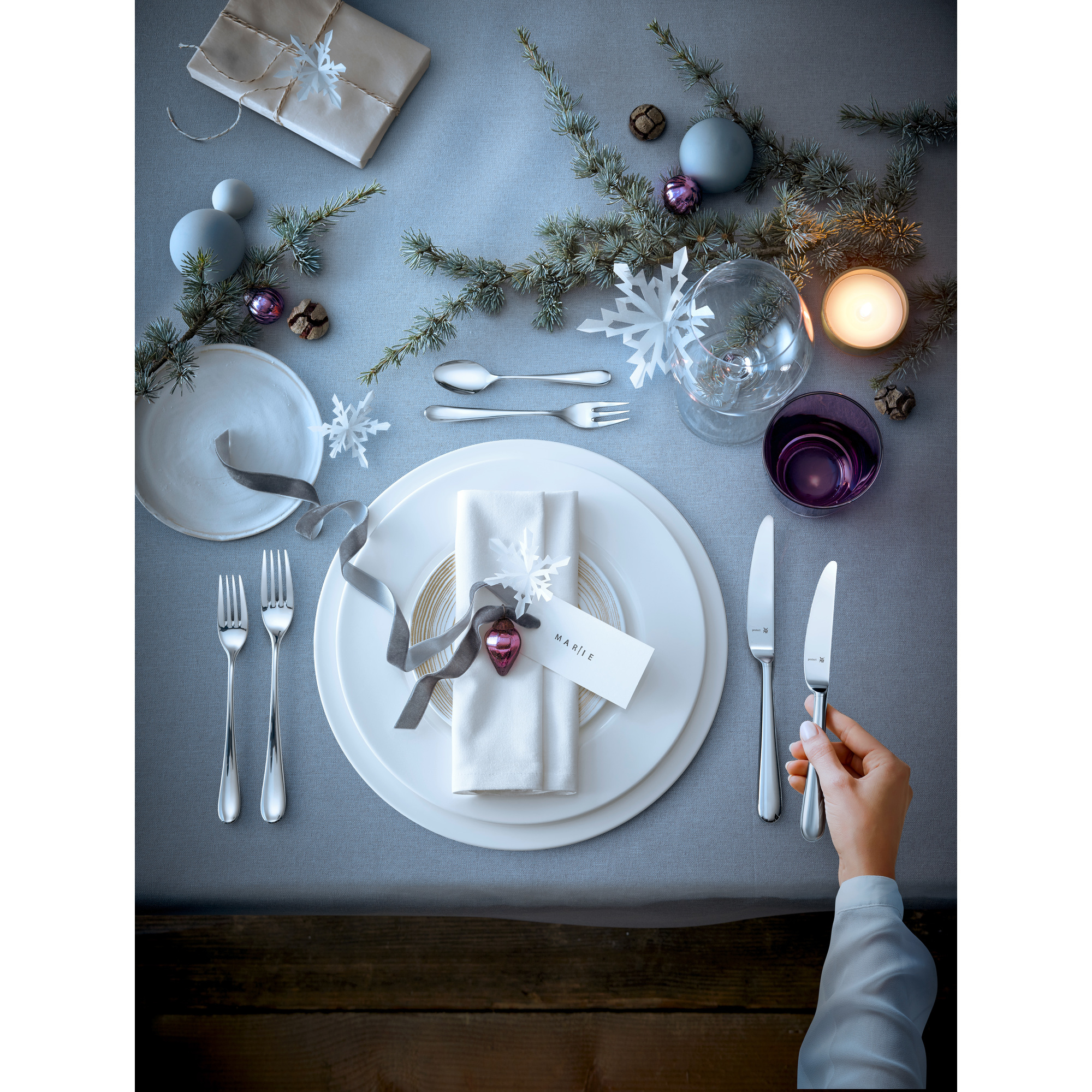 Festliche Tischdekoration mit weißen Tellern, Besteck, Kerze, Ornamenten und Geschenkbox auf grauer Tischdecke.