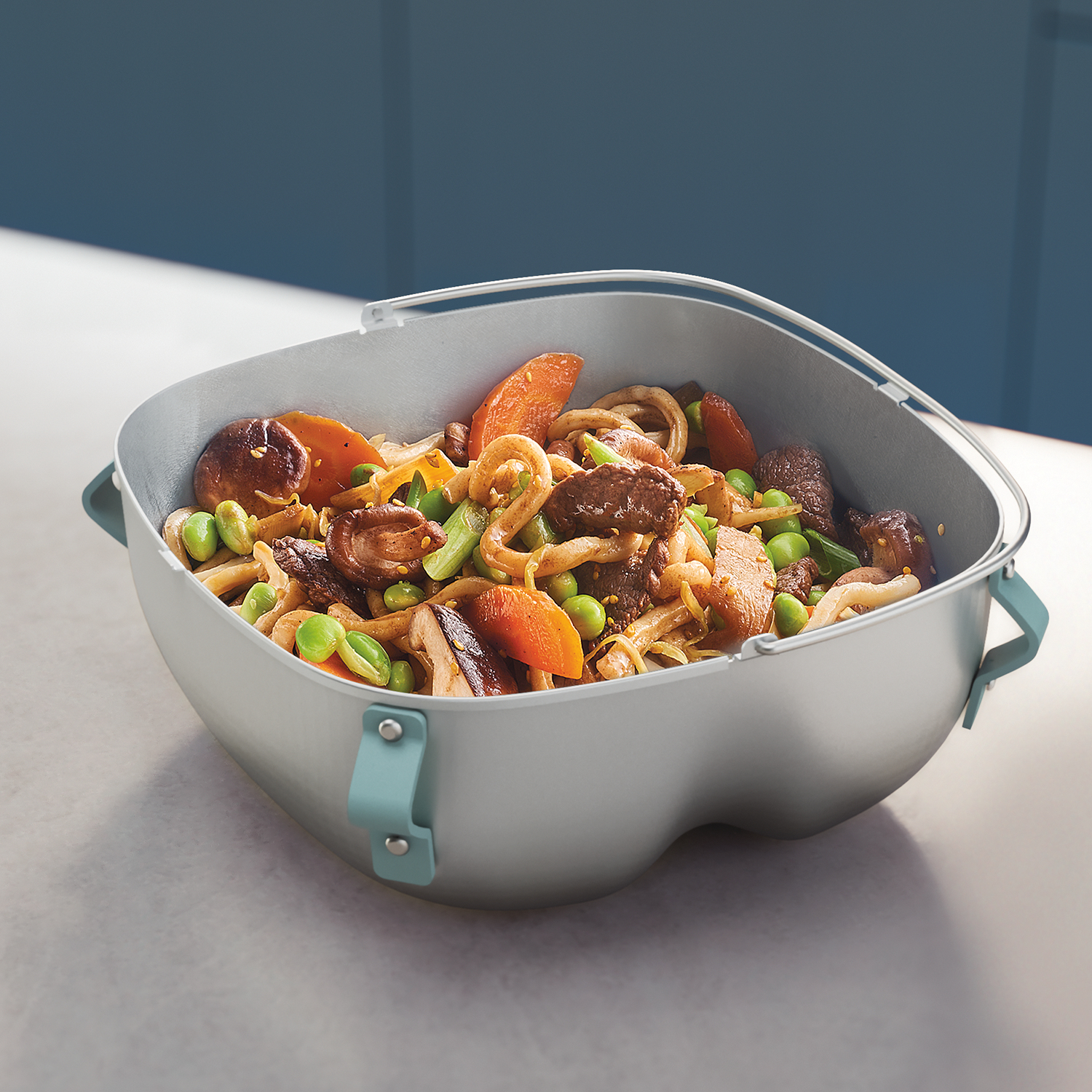 Un contenitore per alimenti grigio pieno di noodles, verdure e carne, su sfondo blu e bianco. Il contenitore ha manici blu.