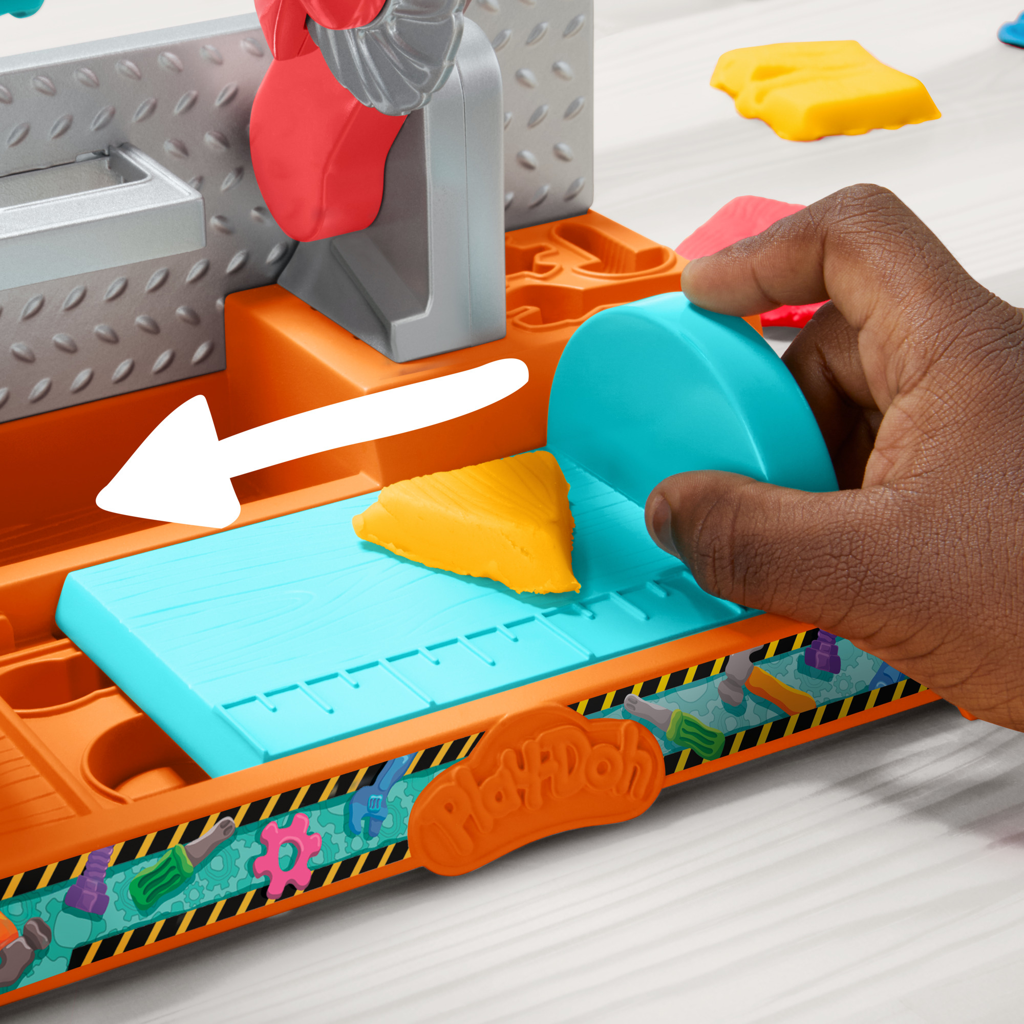 Una mano usa un cortador azul sobre una pieza de Play-Doh amarilla. El juego es naranja, con fondo gris.