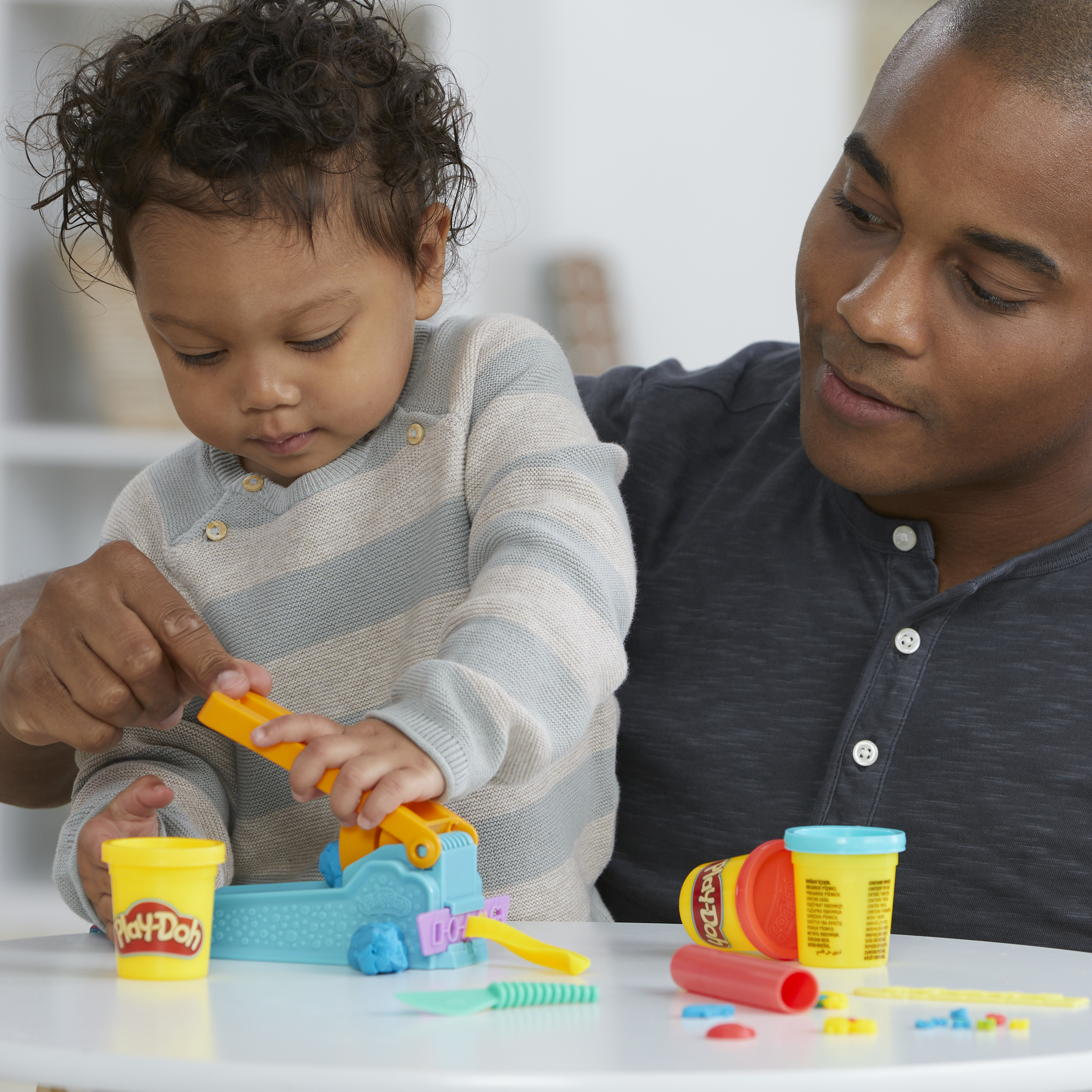Un hombre y un niño juegan con Play-Doh. El niño usa una máquina. Varios recipientes de Play-Doh son visibles.
