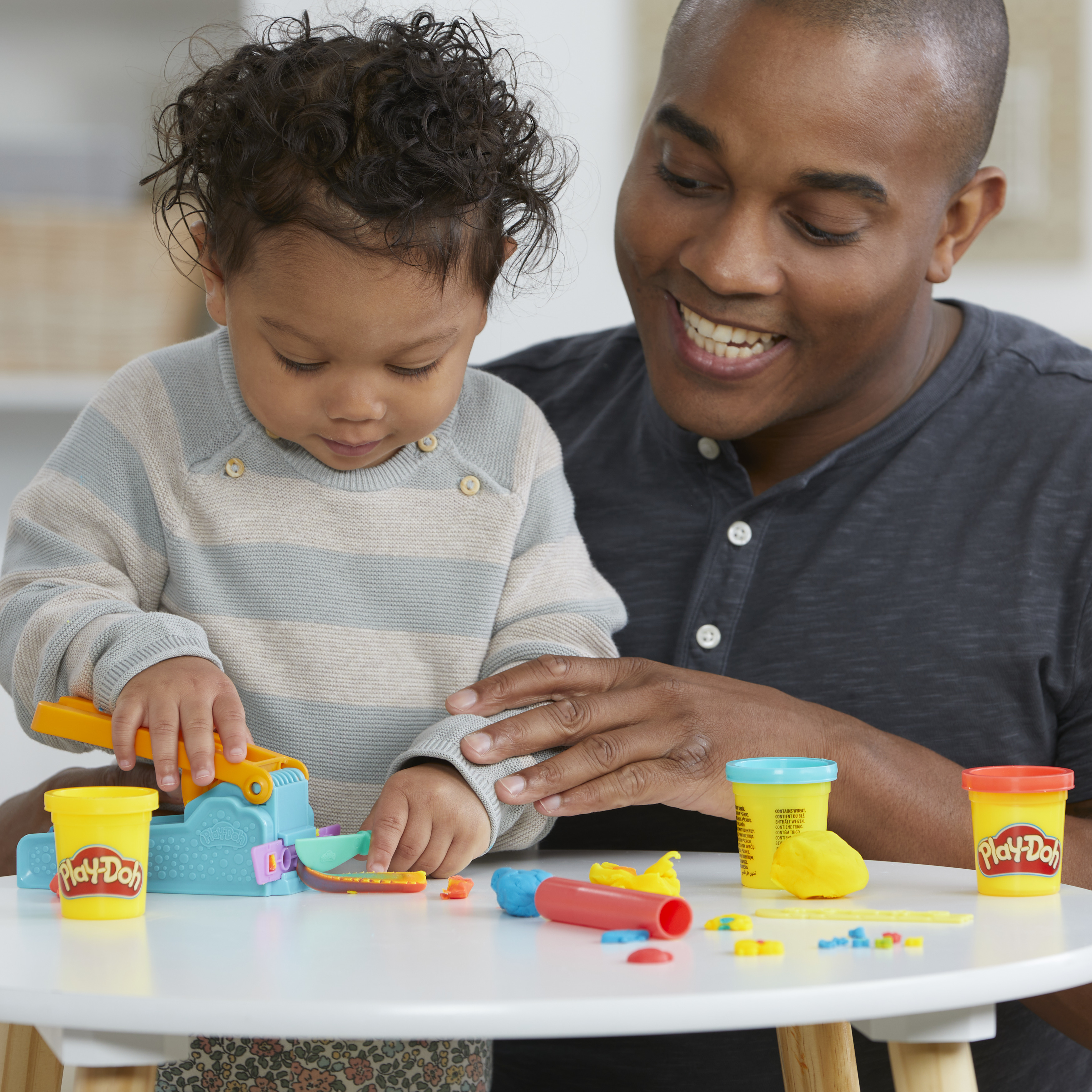 Un niño y un adulto juegan con Play-Doh. El niño usa un juguete. El adulto está sonriendo.