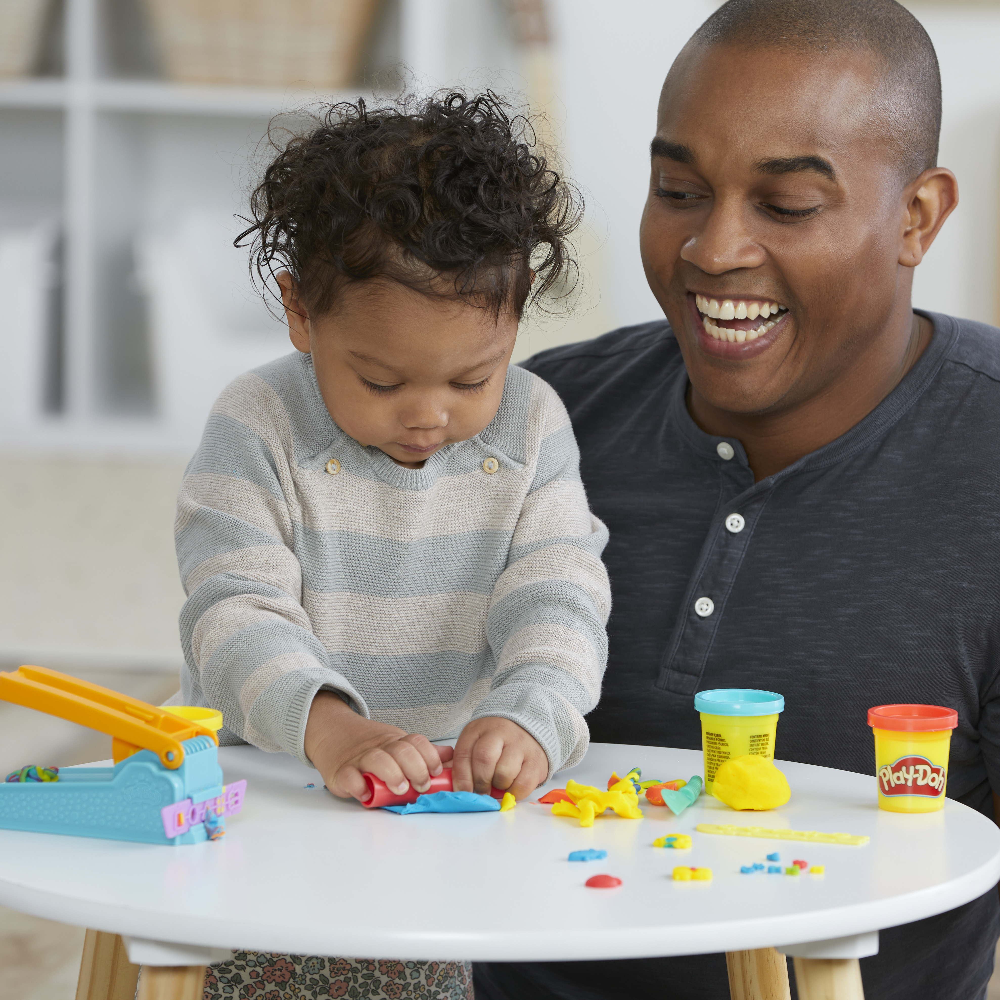 Un hombre y un niño juegan con Play-Doh colorido en una mesa blanca. El hombre sonríe.