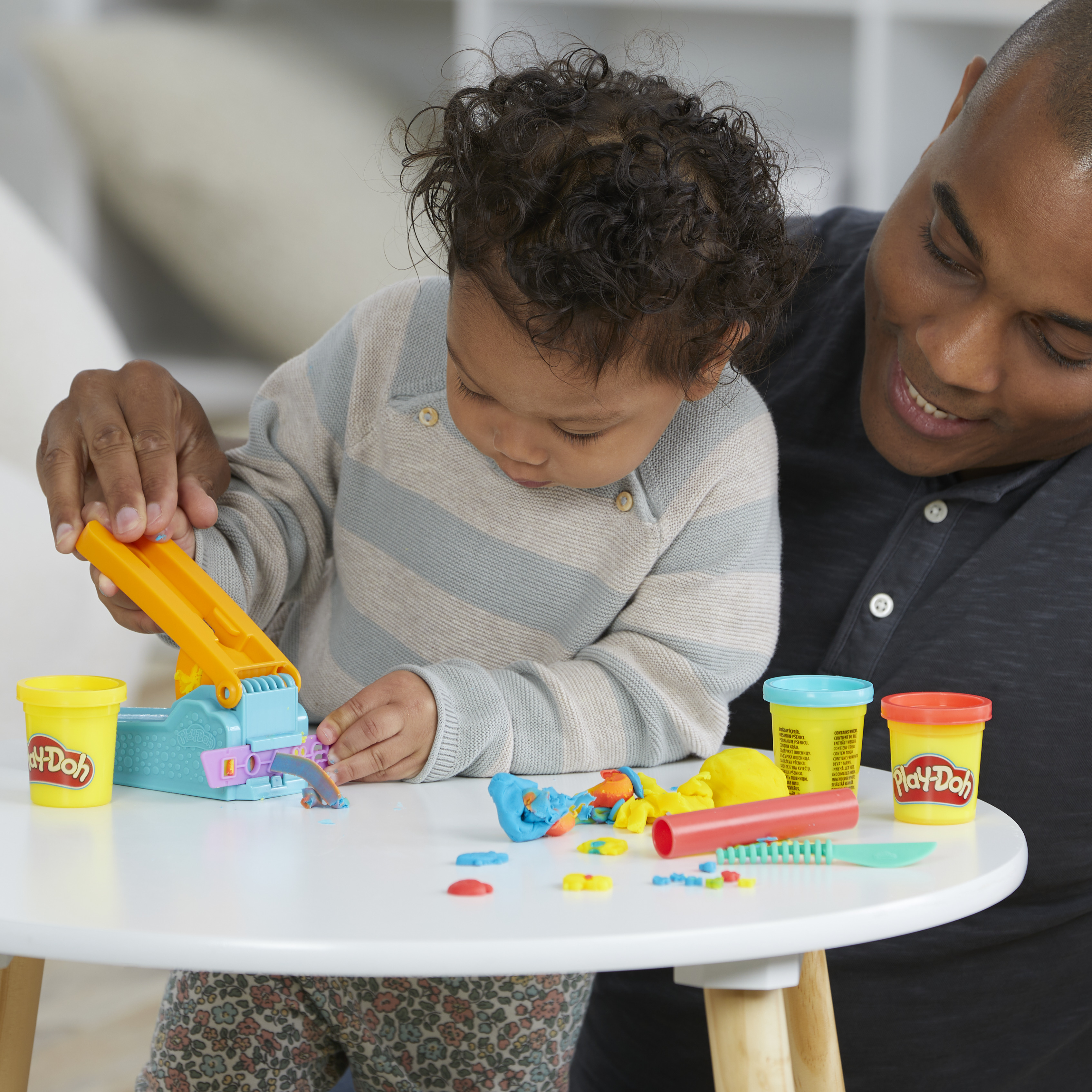 Un hombre y un niño juegan con Play-Doh. El niño usa una prensa de juguete, rodeado de Play-Doh colorido.