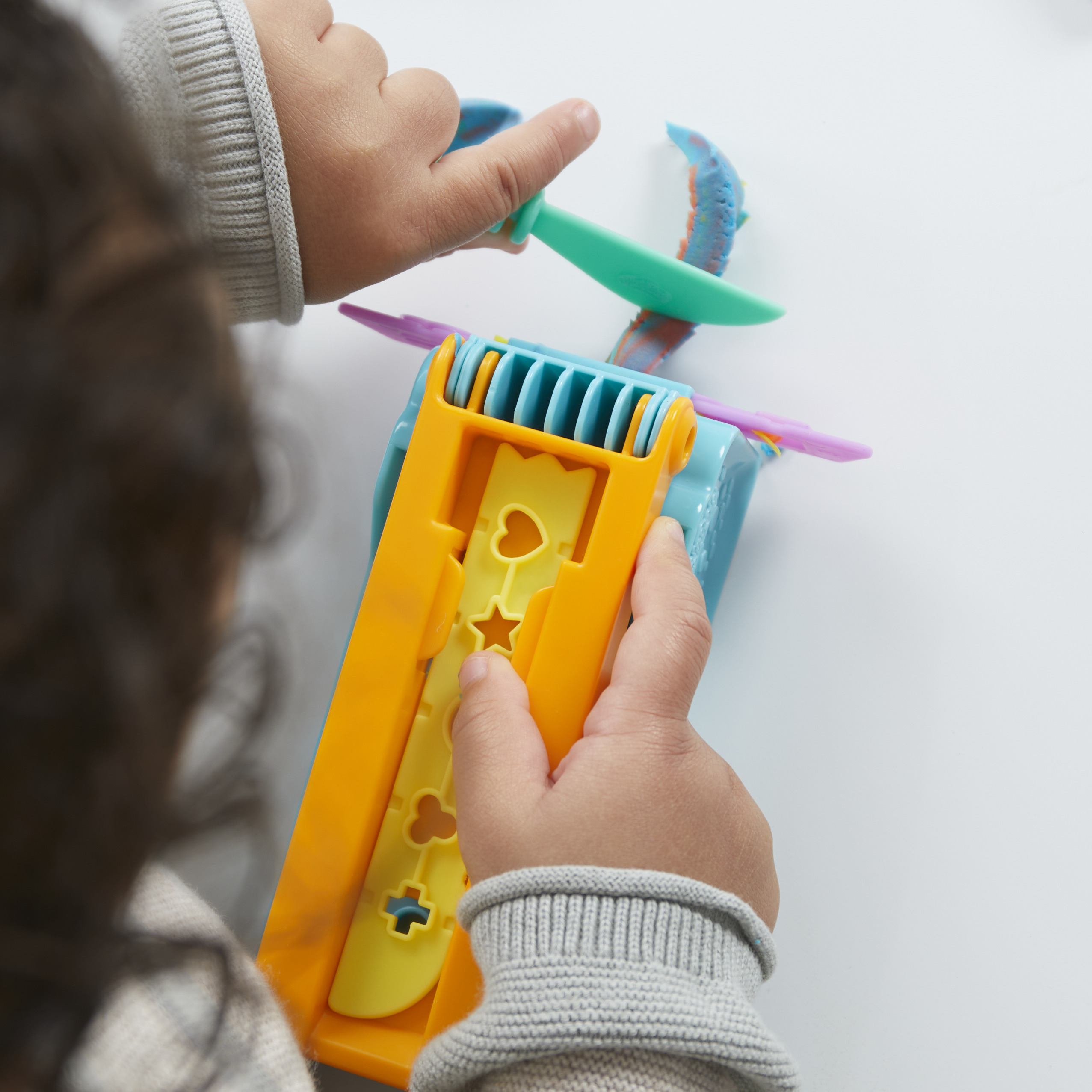 Niño jugando con plastilina y herramientas de colores. Manos visibles, sosteniendo una herramienta y la masa. Fondo blanco.