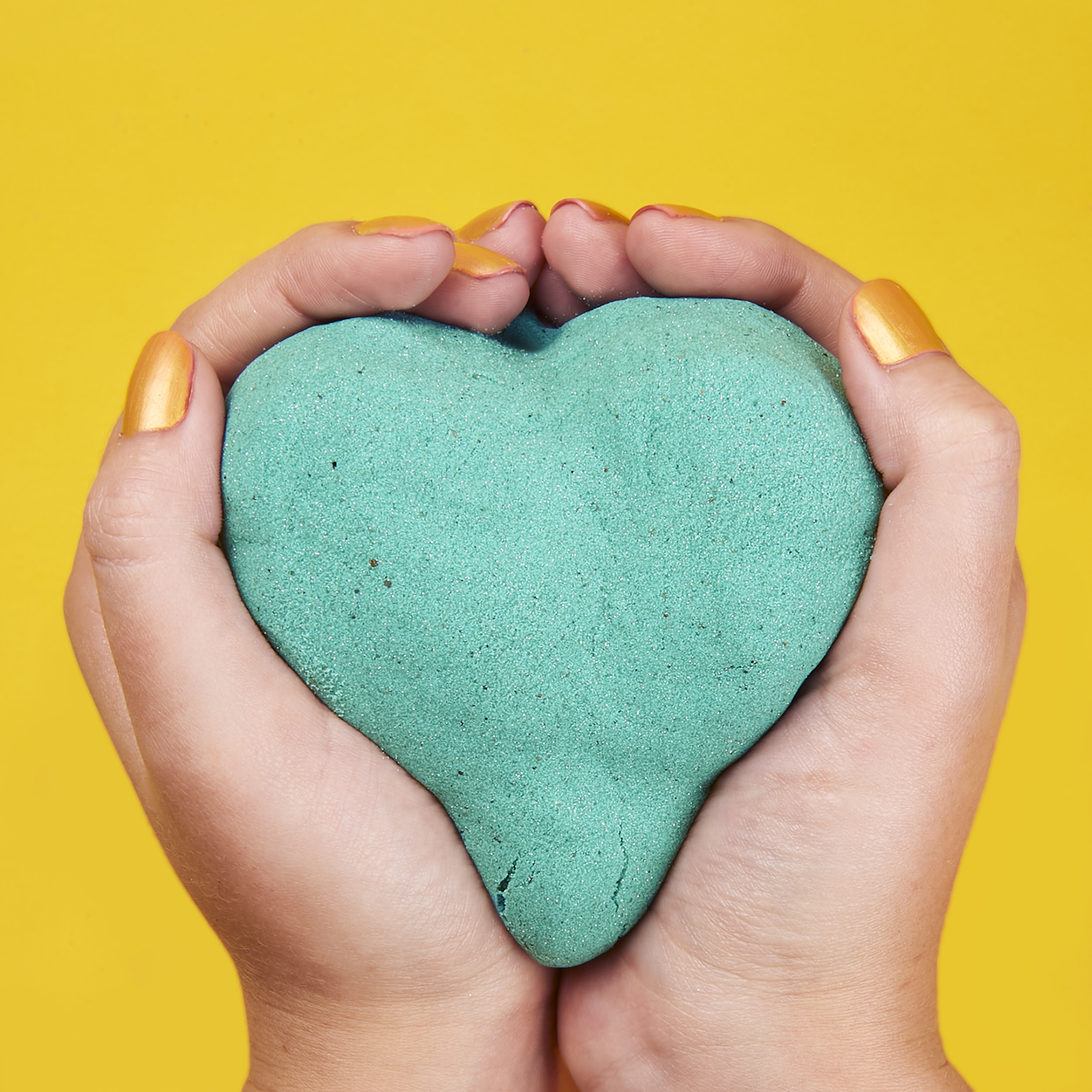 Manos sosteniendo un objeto en forma de corazón, turquesa, contra un fondo amarillo. Uñas doradas.