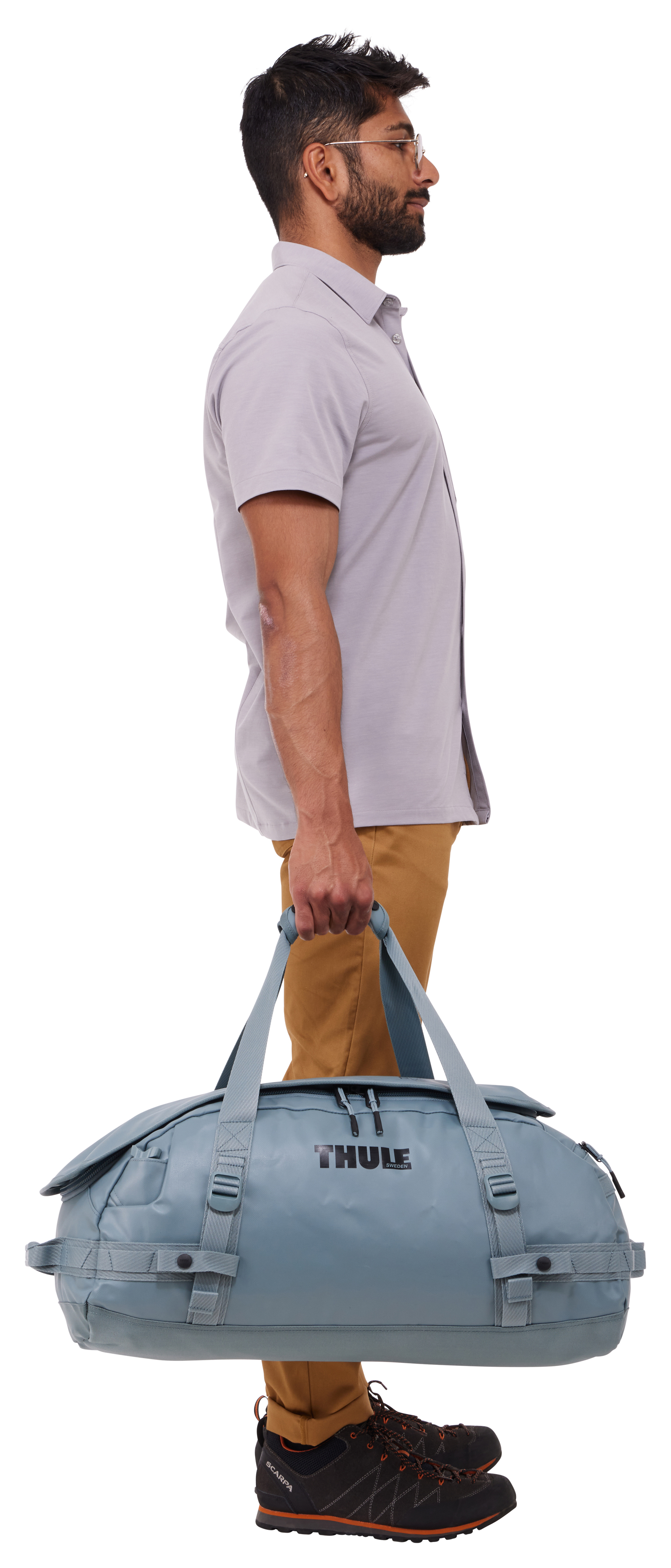 Mężczyzna w szarej koszuli i brązowych spodniach, trzymający niebieską torbę podróżną. W okularach, w profilu na białym tle.