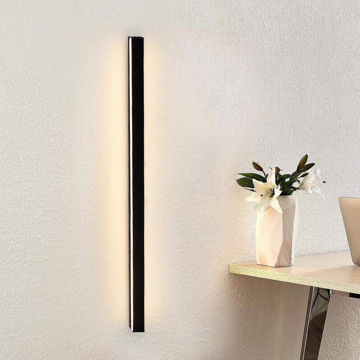 Una moderna lampada da parete è illuminata accanto a un vaso di fiori su un tavolo. Il muro e il tavolo sono chiari.