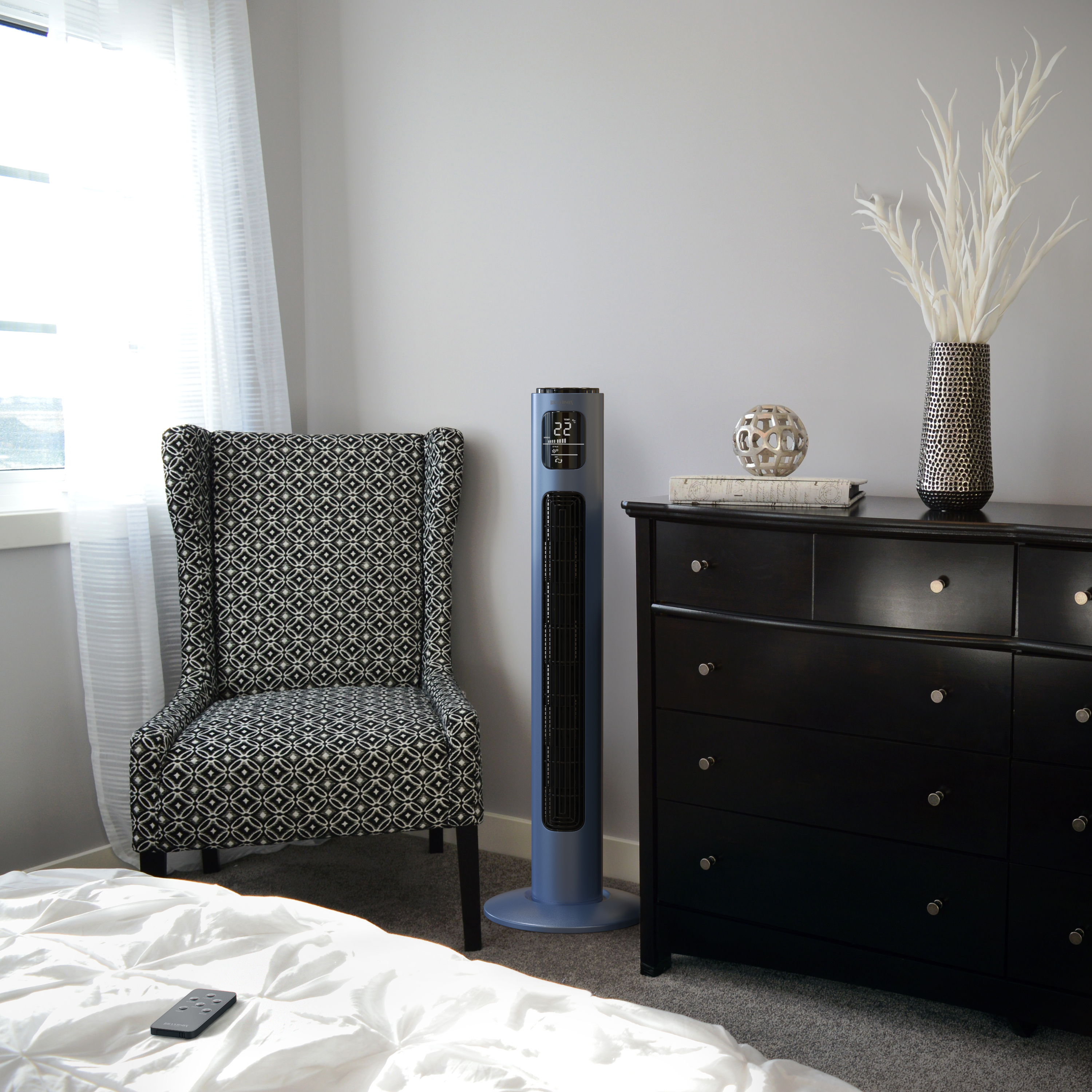Schlafzimmer mit Stuhl, Ventilator, Kommode und Bett. Stuhl ist gemustert, Ventilator blau, Kommode schwarz. Bett mit Fernbedienung.