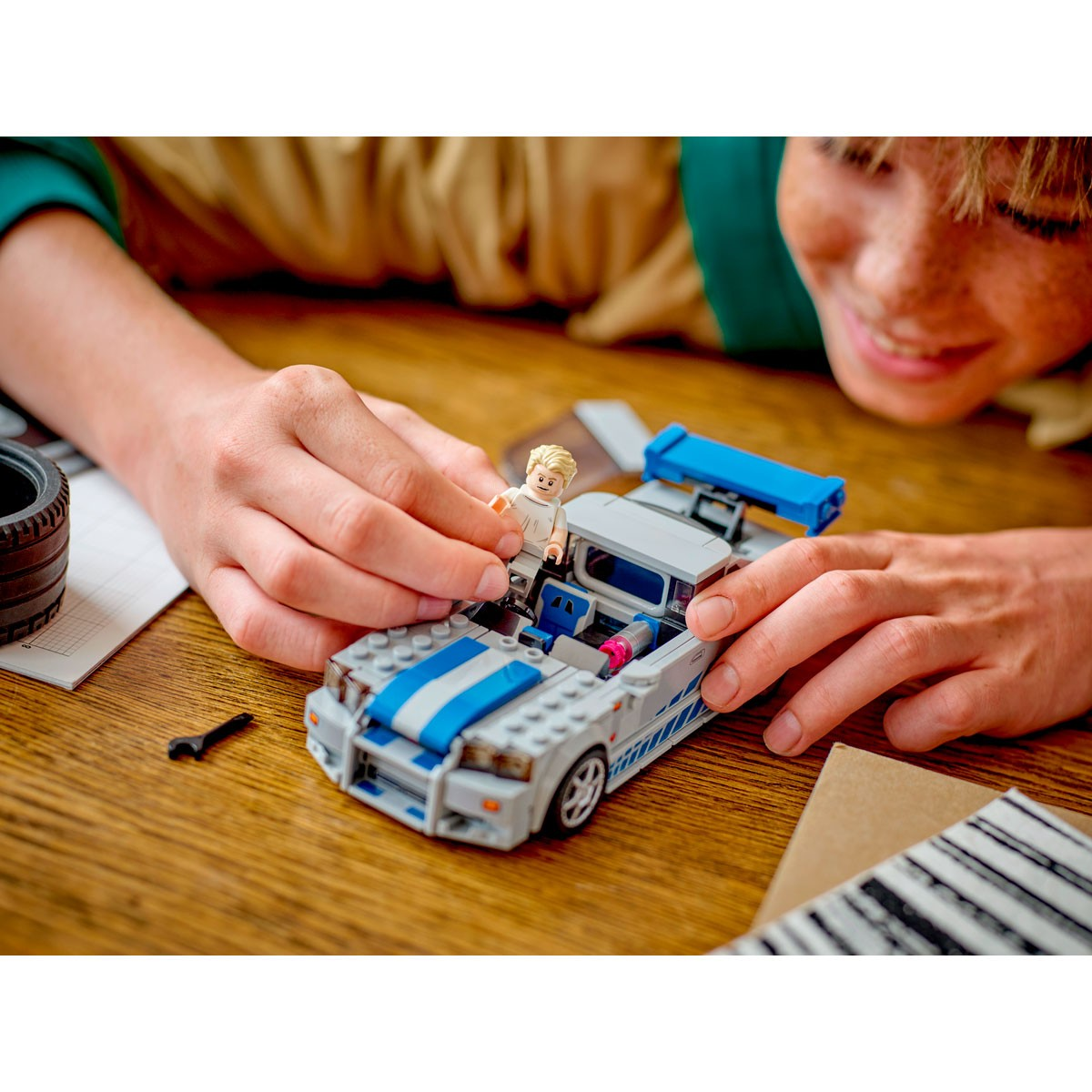 Une personne assemble une voiture Lego. La voiture Lego est grise, avec des rayures bleues. Une figurine Lego est placée à l'intérieur de la voiture.