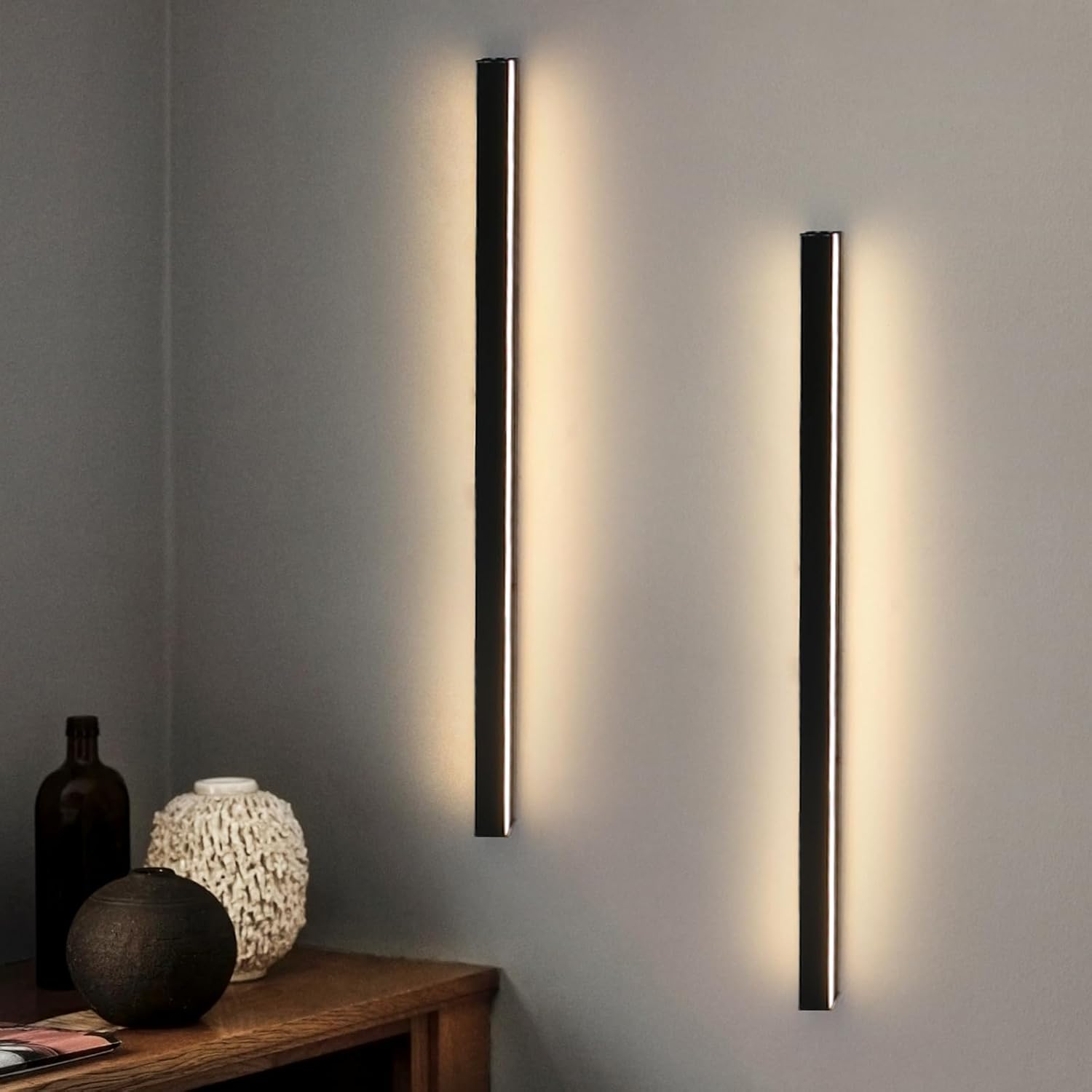 Due lampade da parete nere con una luce calda, accanto a un tavolo di legno con vasi e bottiglie contro un muro grigio.