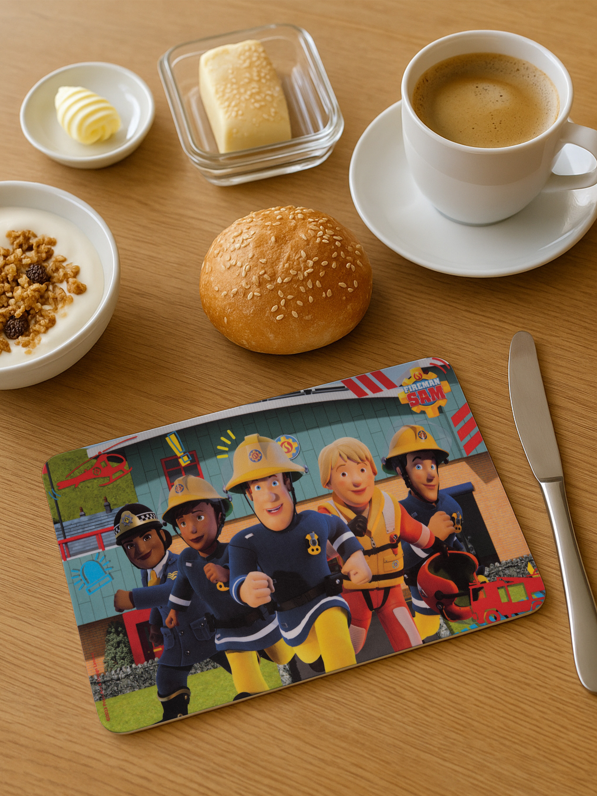 Frühstücksszene: Kaffee, Butter, Joghurt mit Müsli, Brötchen und eine Fireman Sam-Platzmatte auf einem Holztisch.