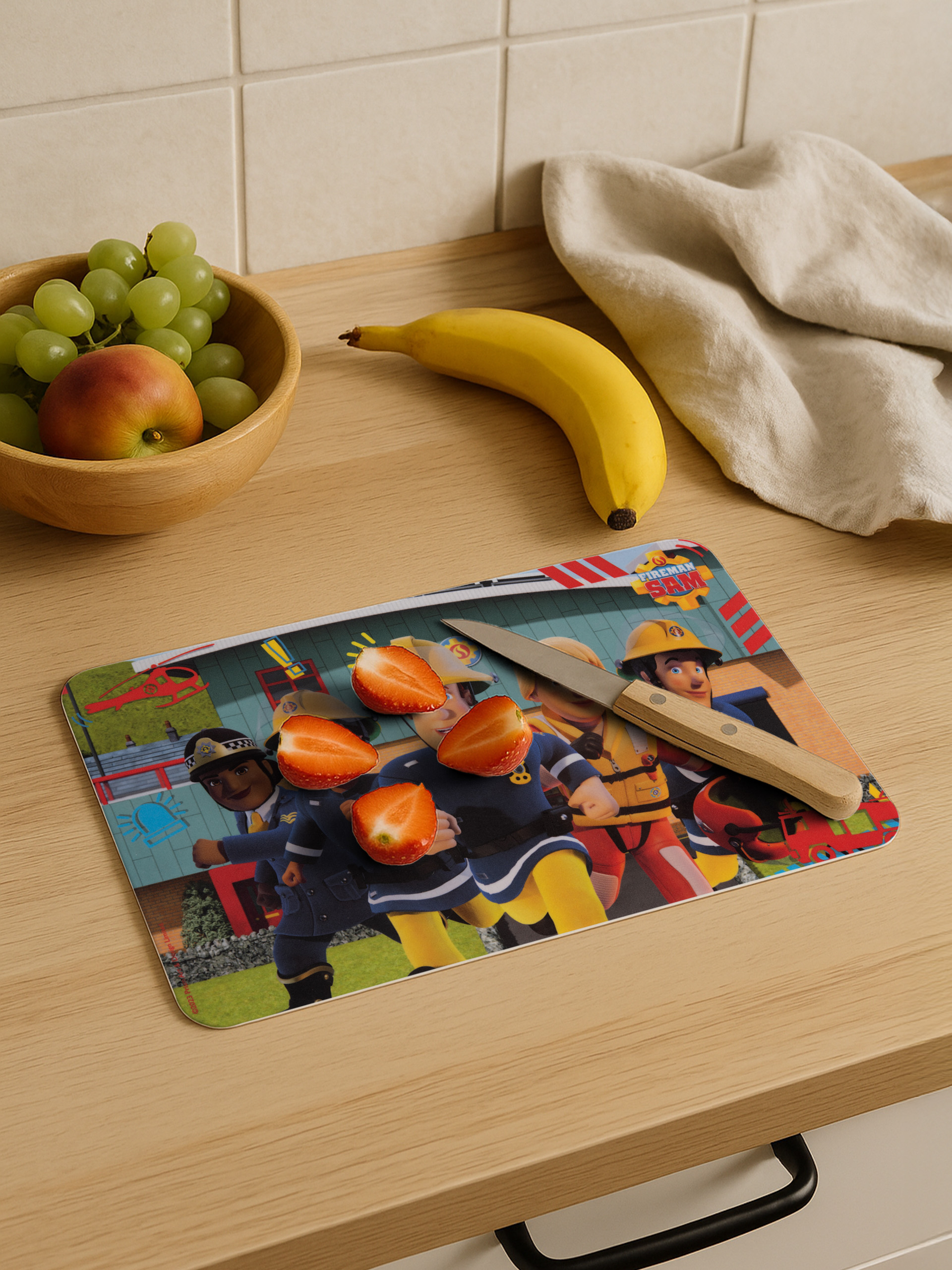 Ein Schneidebrett mit Feuerwehrmann Sam-Grafiken, Erdbeeren, ein Messer, eine Banane, Trauben und ein Apfel auf Holz.