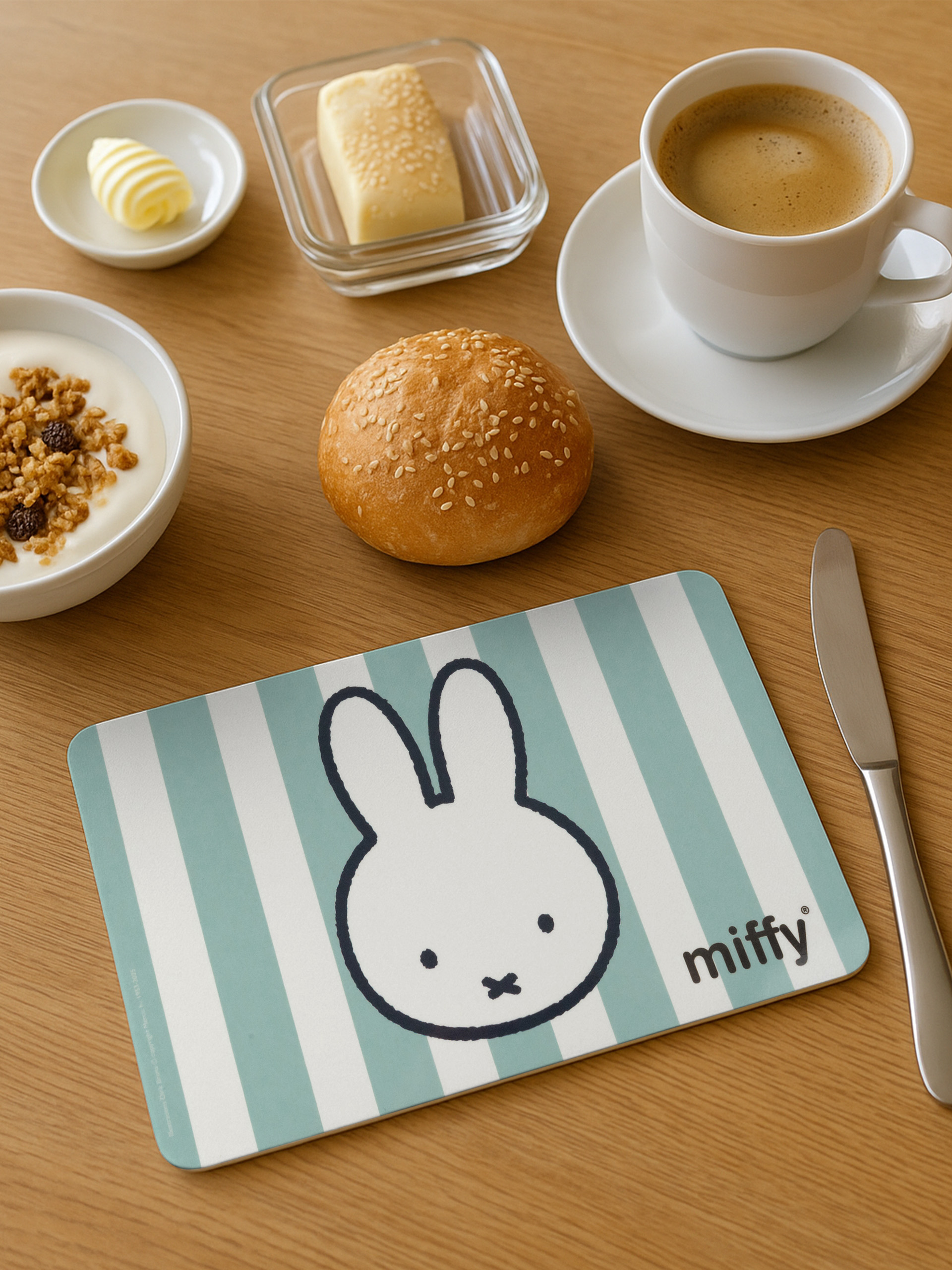 Frühstücksszene: Kaffee, Brötchen, Butter, Joghurt, Miffy-Tischset. Holztisch. Draufsicht.