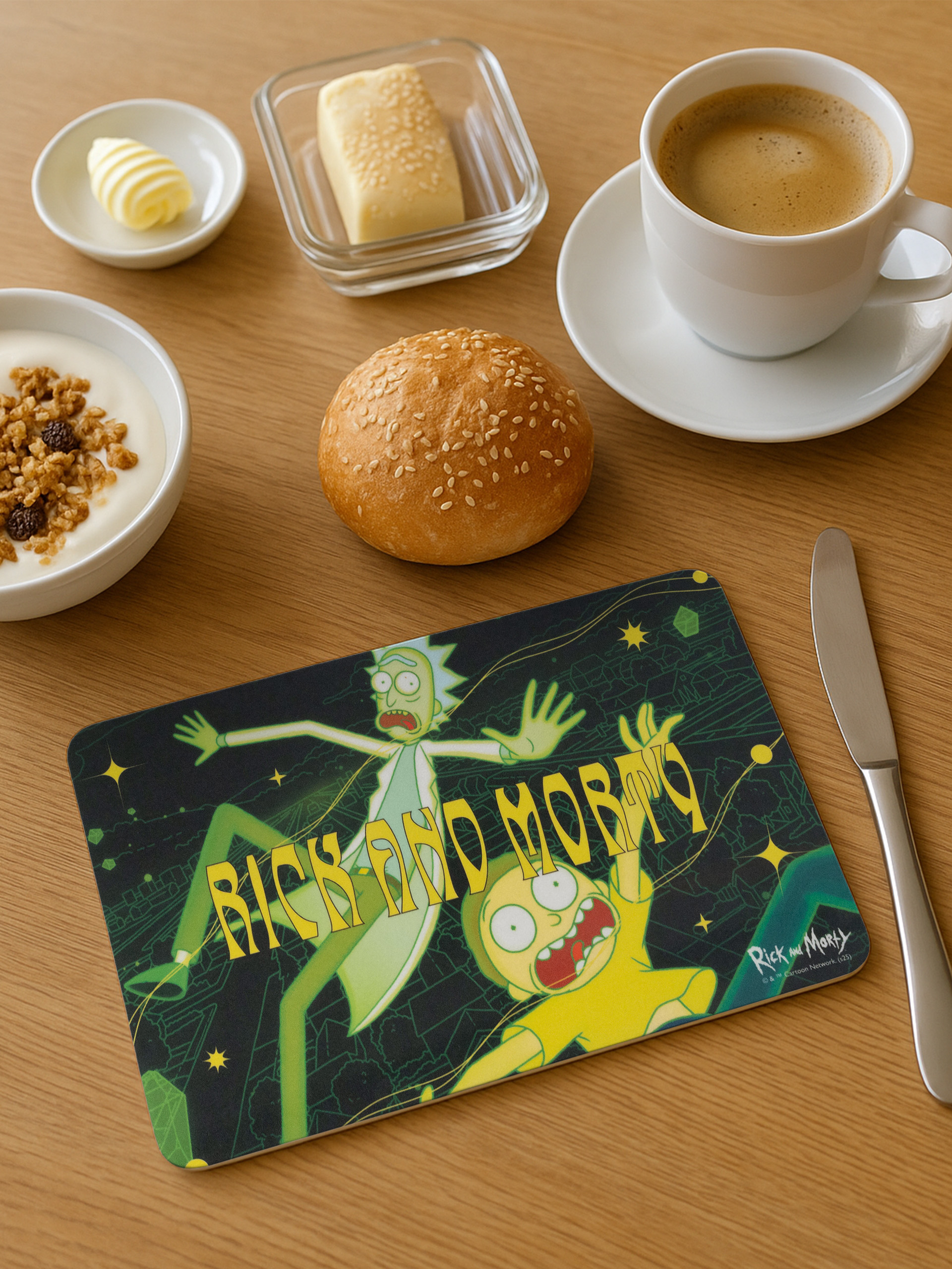 Frühstücksszene mit Rick-und-Morty-Untersetzer, Brot, Joghurt, Butter, Kaffee und Messer auf Holztisch.