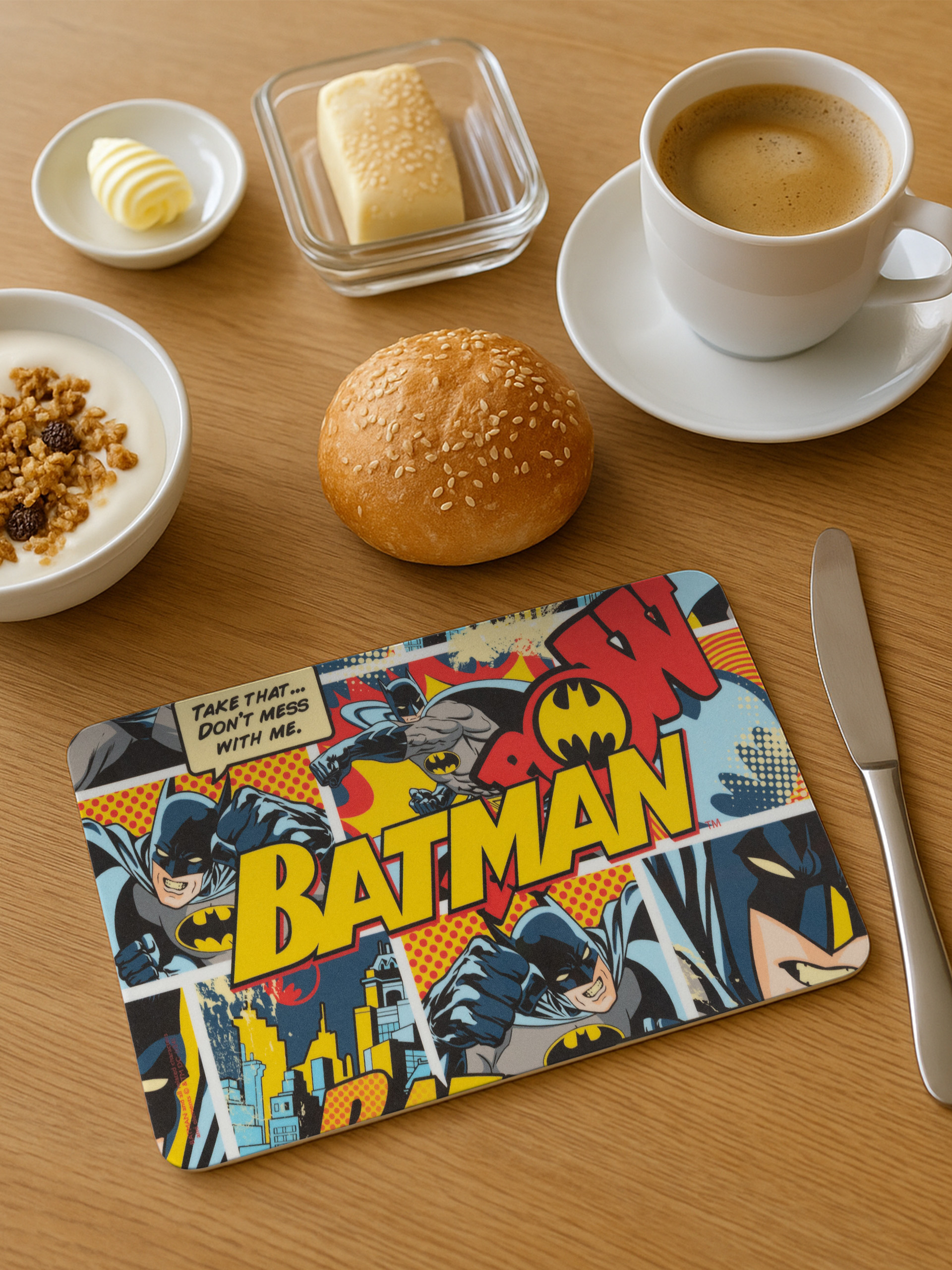 Frühstücksszene: Kaffee, Butter, Joghurt, Brötchen und eine Batman-Tischmatte auf einem Holztisch.