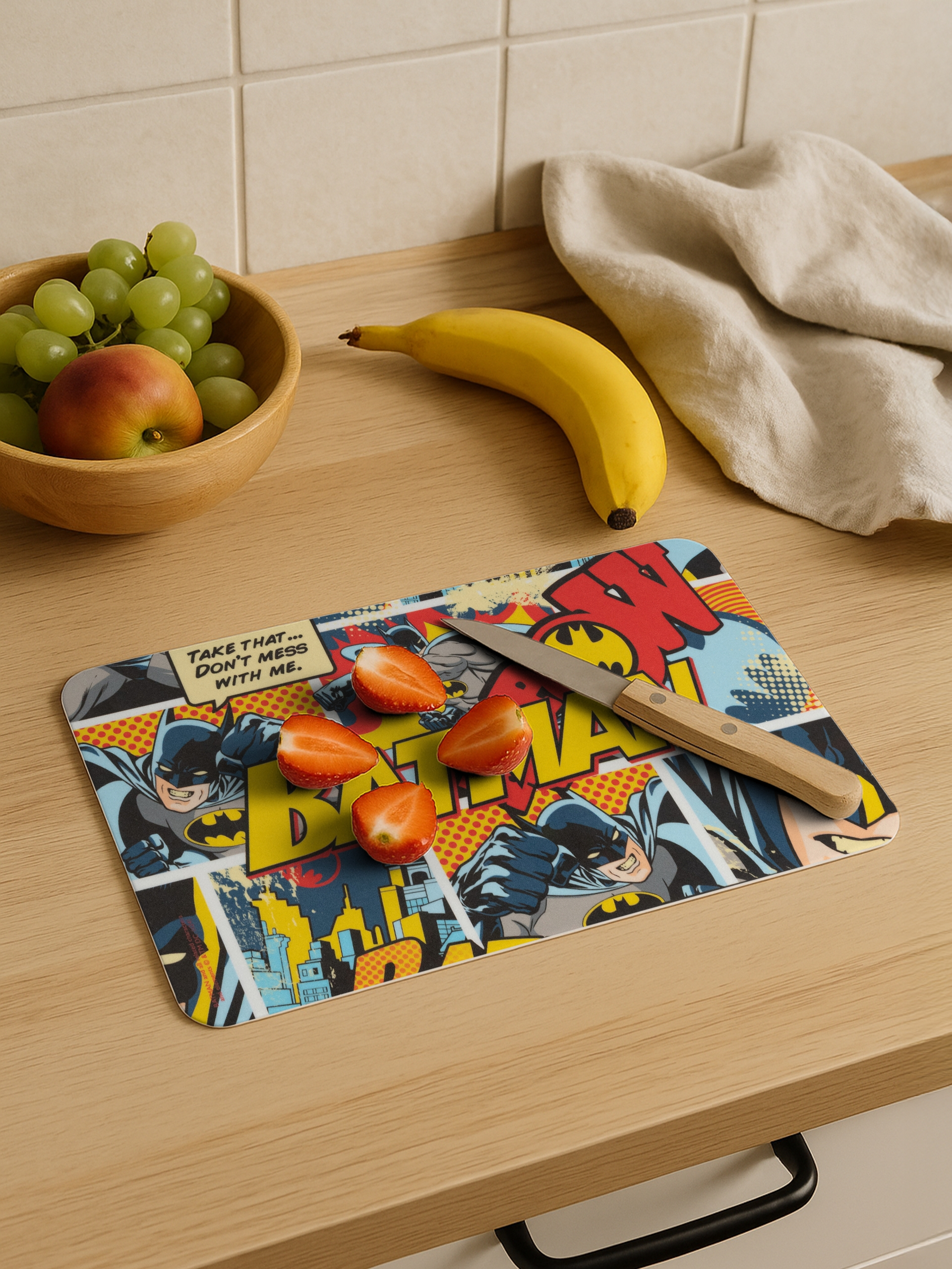 Ein Batman-Schneidebrett mit Erdbeeren und einem Messer, mit Banane, Trauben und Apfel daneben.