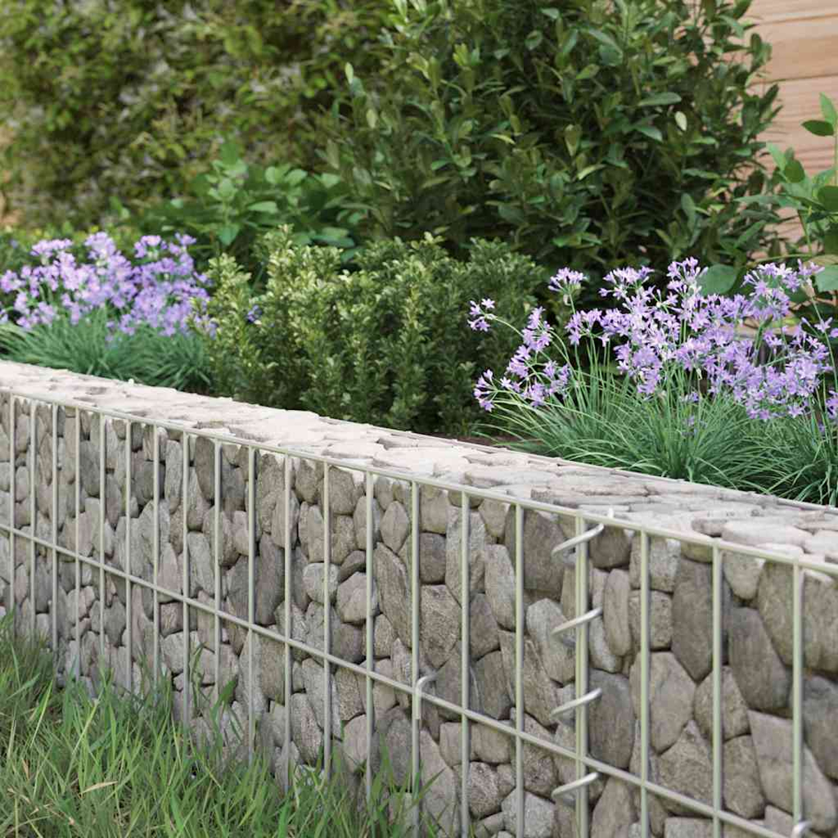 Eine Steinmauer mit Drahtzaun, lila Blumen, grüne Büsche und Gras in einem Garten.