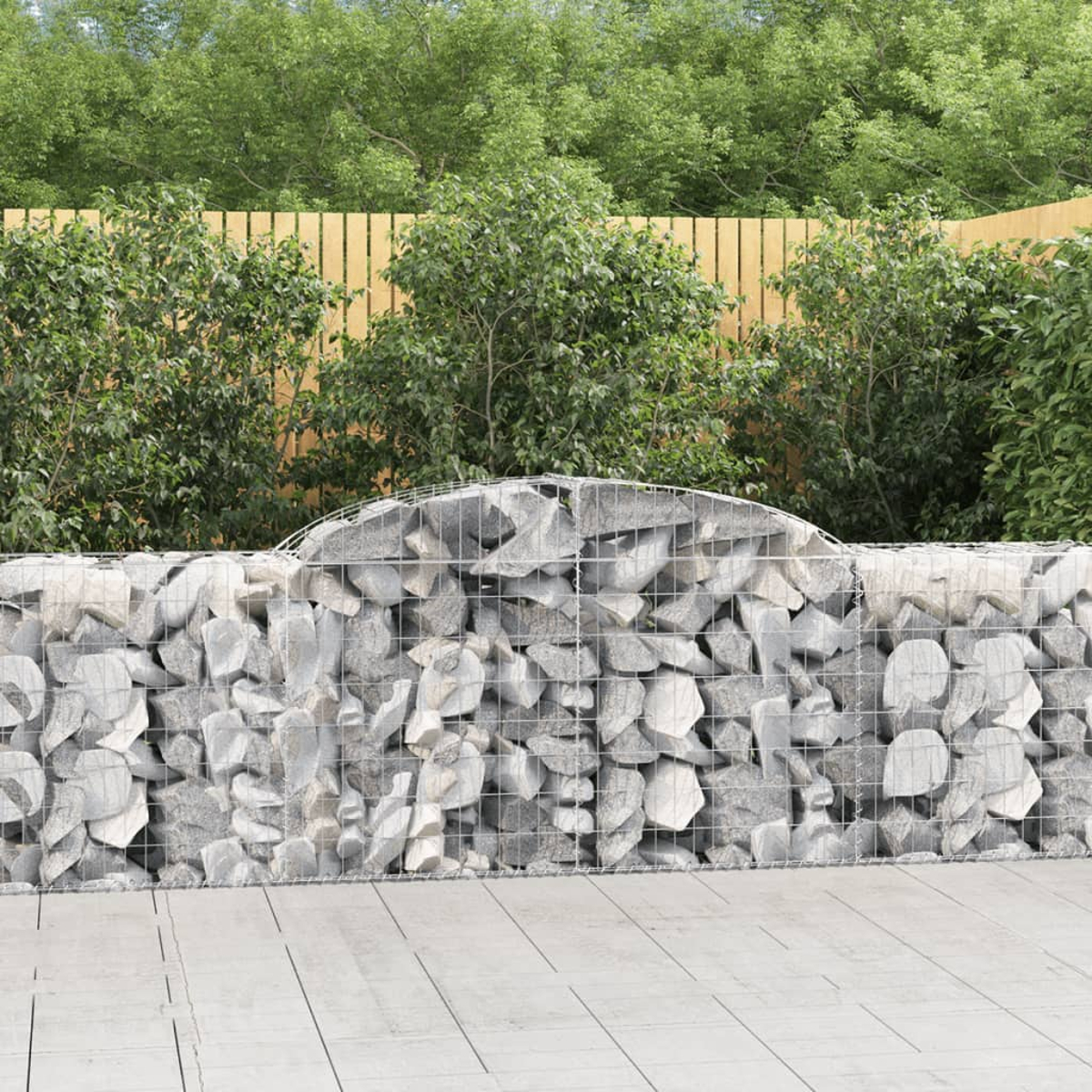 Eine Steinmauer im Garten. Im Hintergrund grüne Büsche und ein Holzzaun. Im Vordergrund eine graue Stein-Terrasse.