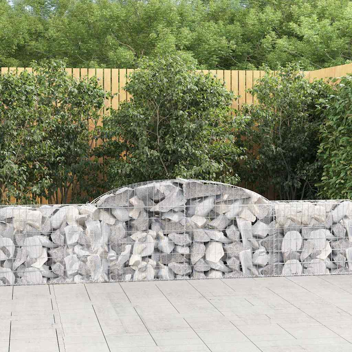 Eine Stein-Gabionenwand, gefüllt mit grauen Steinen, steht vor einem Holzzaun und grünen Büschen.