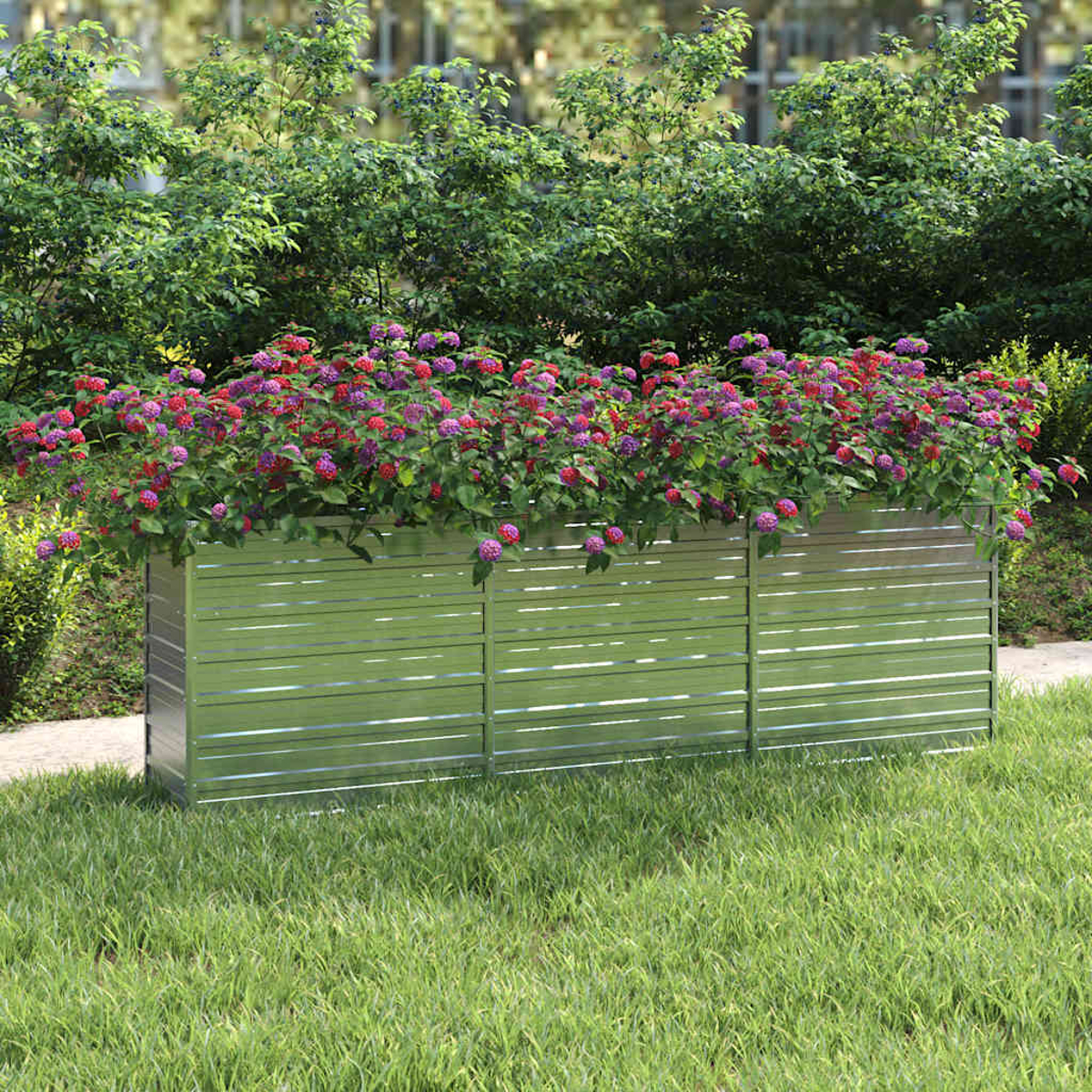 Eine Metallpflanzkasten mit bunten Blumen steht auf Gras. Büsche und Bäume bilden einen grünen Hintergrund.