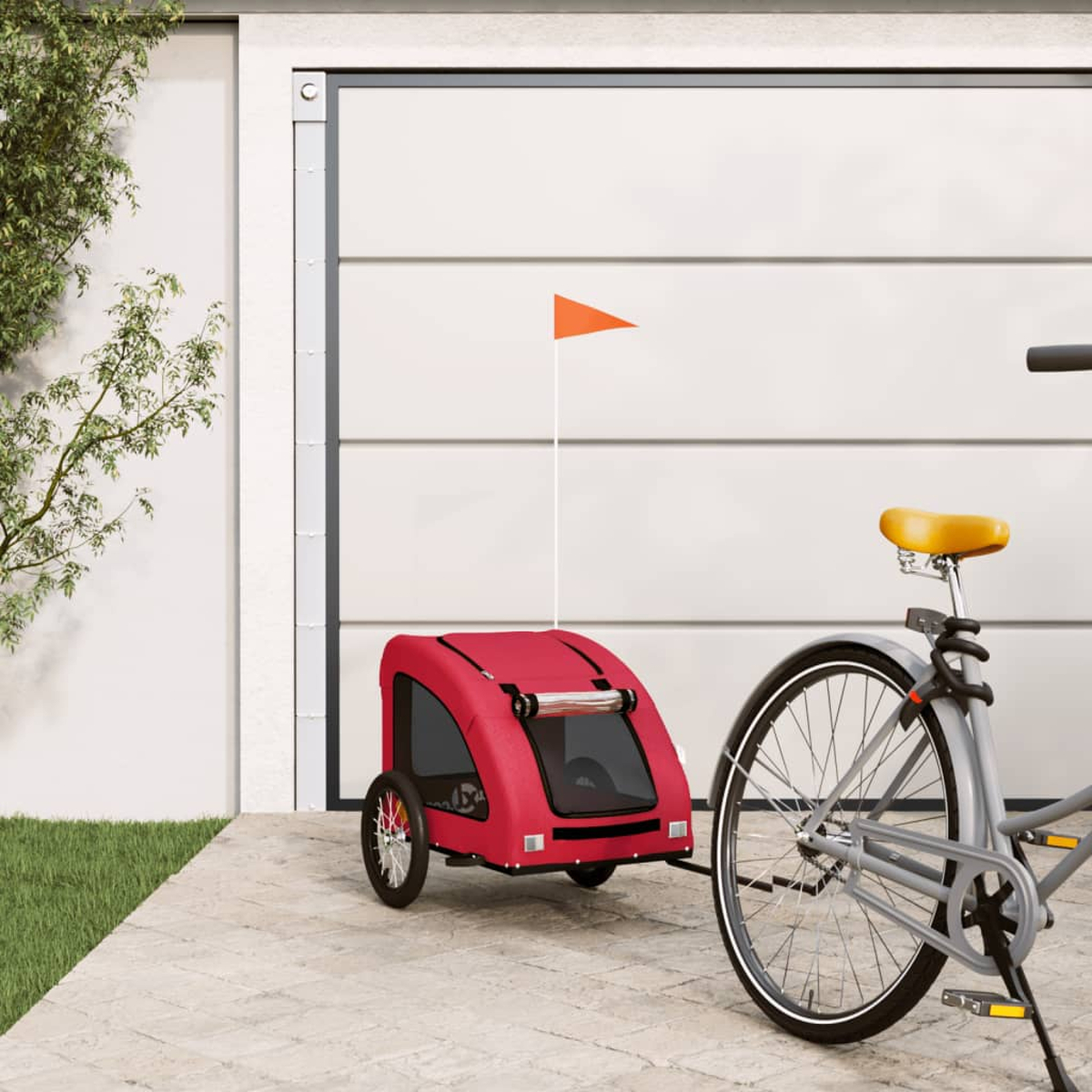 Ein roter Fahrradanhänger mit einer orangefarbenen Flagge ist an einem grauen Fahrrad befestigt. Im Hintergrund eine weiße Garagentür und grünes Gras.