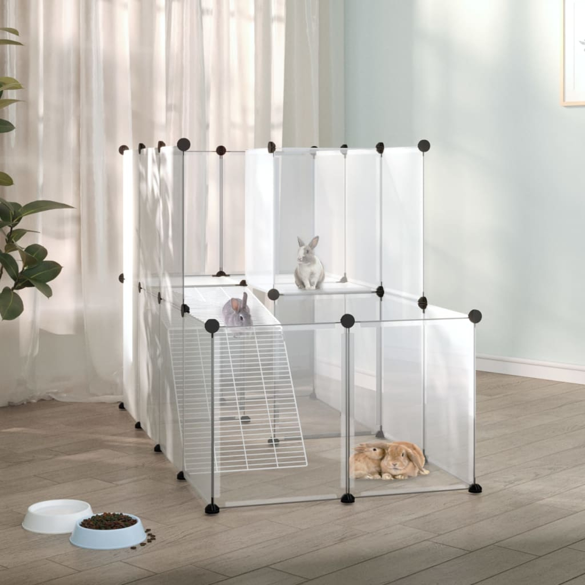 Drei Kaninchen in einem transparenten Kunststoffgehege. Zwei Schalen stehen davor. Weiße Vorhänge und eine Pflanze im Hintergrund.
