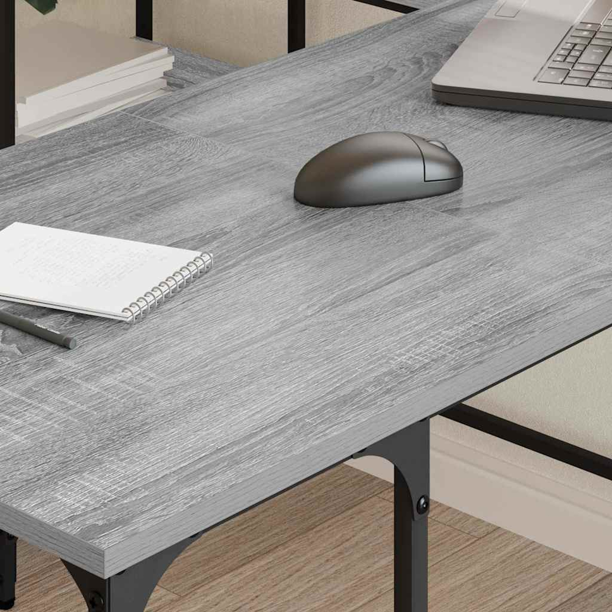 Ein grauer Holzschreibtisch mit Laptop, Maus, Notizblock und Stift. Der Schreibtisch hat schwarze Metallbeine. Hintergrund ist eine Wand und ein Regal.