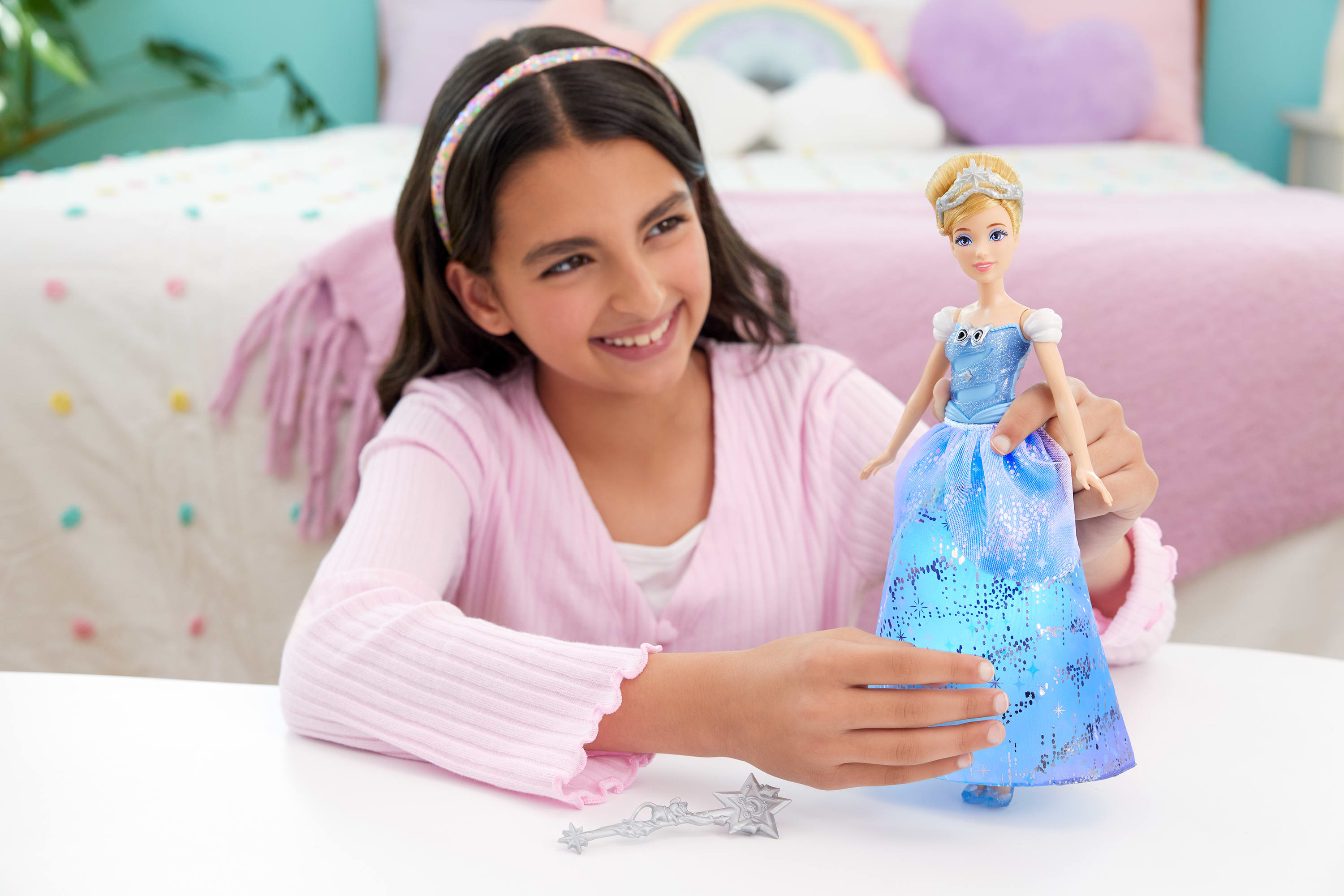Ein lächelndes Mädchen hält eine Cinderella-Puppe. Ein Zauberstab liegt auf dem Tisch. Im Hintergrund ist ein Bett.