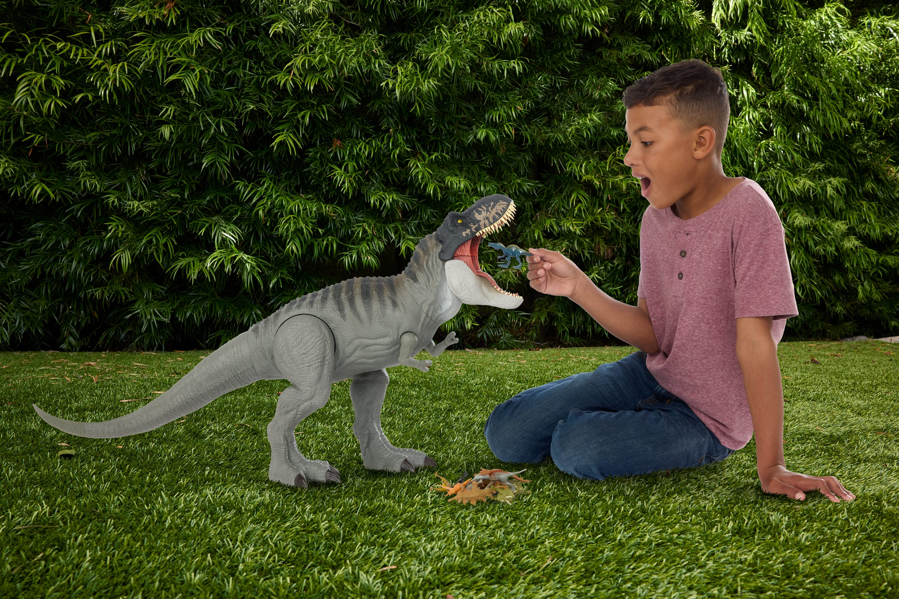 Ein Junge mit einem Dinosaurierspielzeug interagiert mit einem großen Dinosaurierspielzeug. Eine grüne Hecke im Hintergrund.
