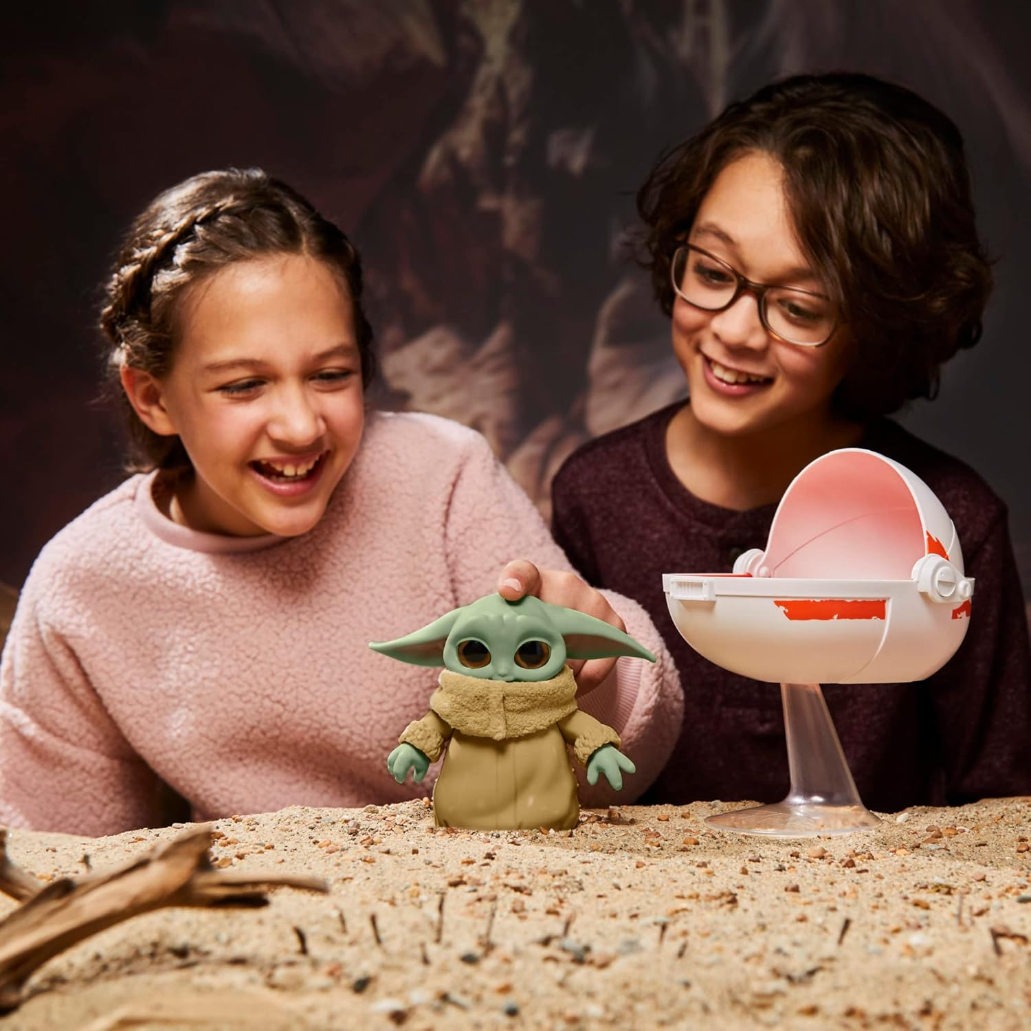 Dos niños sonrientes juegan con un juguete de Grogu y una cuna sobre arena. El fondo es una imagen oscura y borrosa.