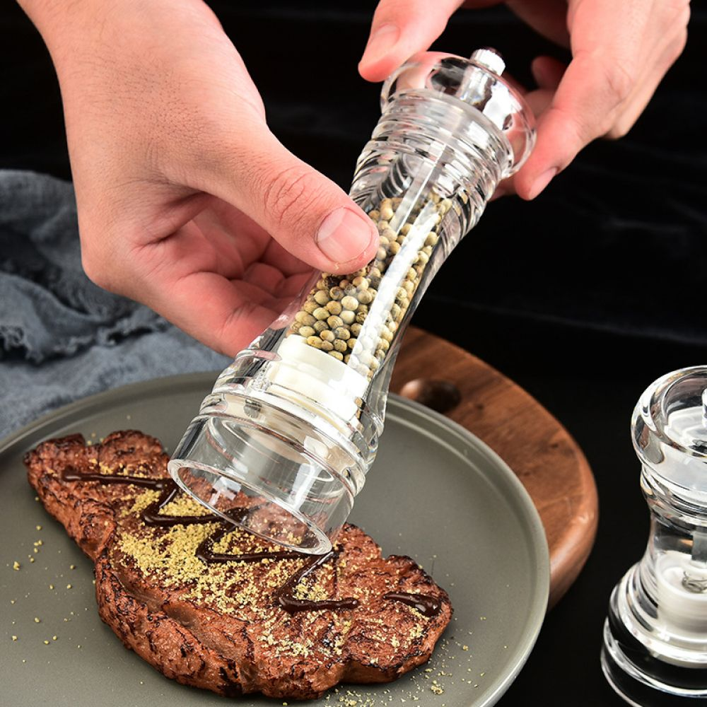 Eine Person mahlt Pfeffer auf ein Steak. Ein klarer Pfeffermühle wird über dem Steak auf einem grauen Teller gehalten. Holzbrett im Hintergrund.