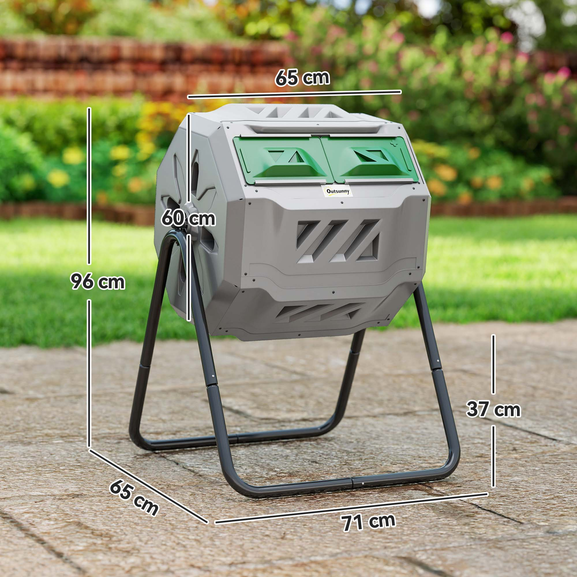 Un composter grigio con coperchi verdi, telaio nero e dimensioni etichettate. Lo sfondo mostra piante verdi e un muro di mattoni.