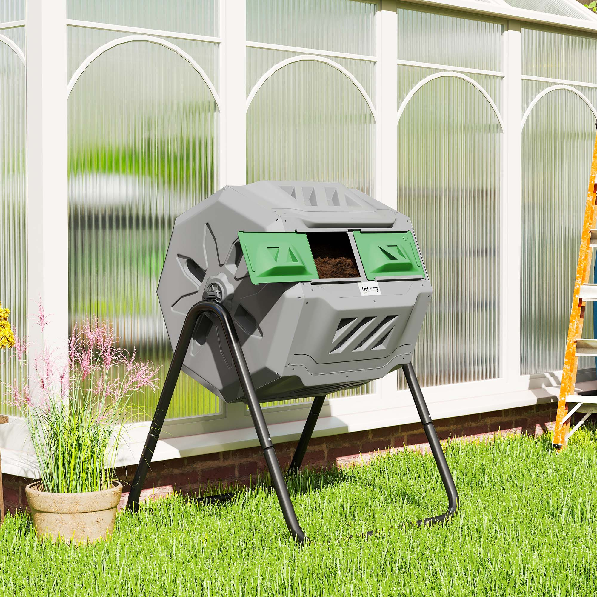 Un composter grigio con coperchi verdi è sull'erba verde. Una serra è sullo sfondo.