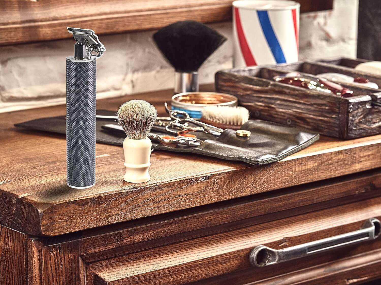 Friseurbedarf auf Holzoberfläche. Enthält Trimmer, Rasierpinsel, Schere und einen Behälter mit rot-weiß-blauen Streifen.