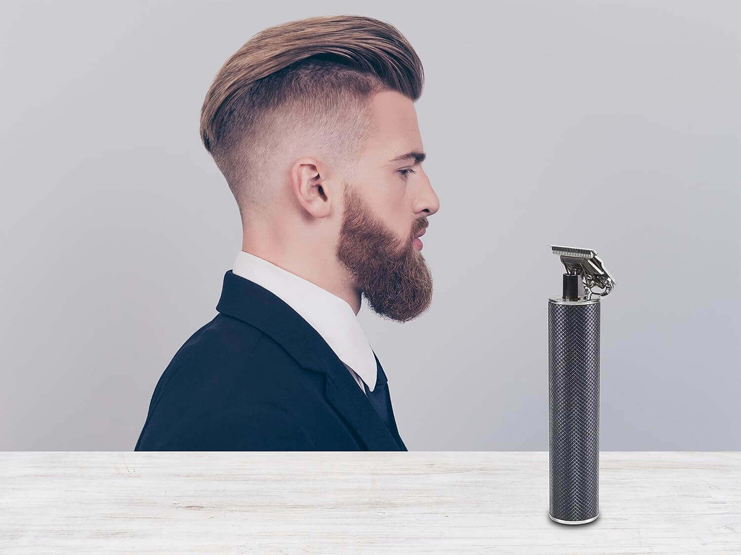 Ein Mann mit Bart und stilvollem Haarschnitt. Ein Trimmer steht neben ihm. Der Mann trägt einen Anzug.