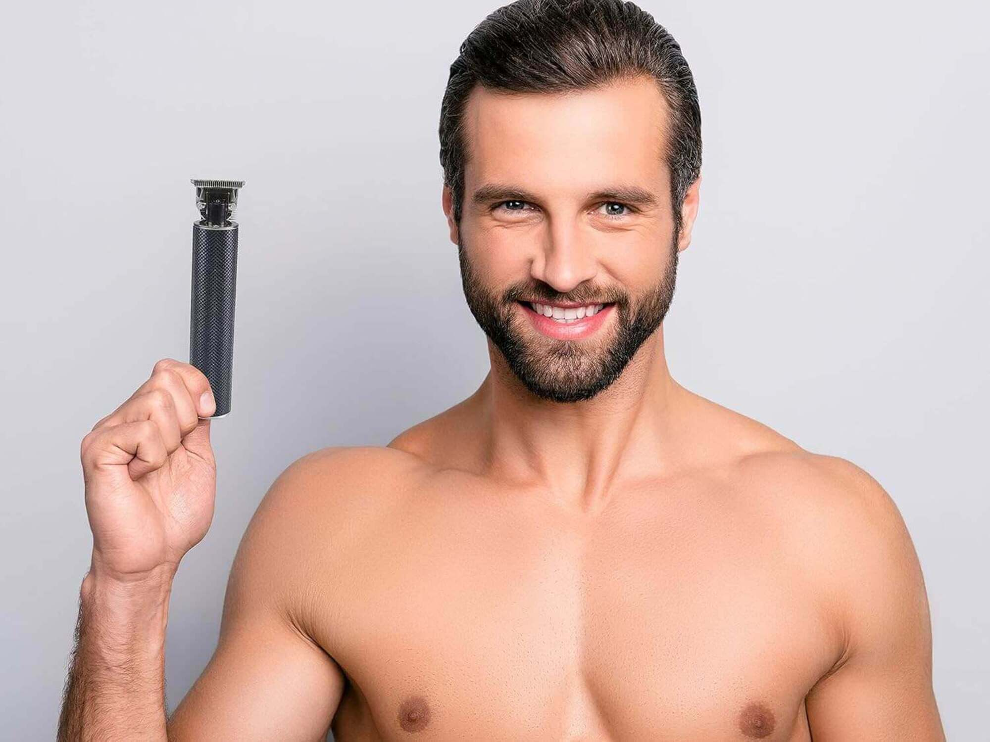 Ein Mann mit Bart hält einen Trimmer und lächelt in die Kamera. Er ist ohne Hemd. Der Hintergrund ist grau.