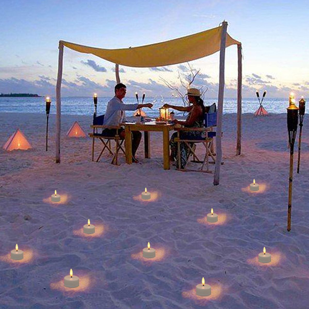 Una coppia cena su una spiaggia al crepuscolo. Candele e torce illuminano la scena. Una tenda da sole ombreggia il tavolo con la coppia.