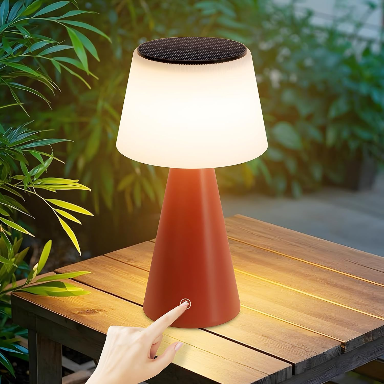 Una mano presiona el botón de una lámpara. La lámpara está sobre una mesa de madera. Follaje verde en el fondo. La lámpara es roja y blanca.